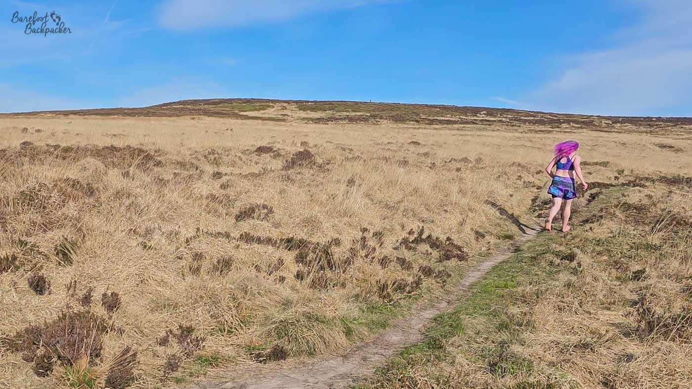 A barefoot runner with bright pink hair follows a narrow path across open heather moorland, surrounded by dry grass and an expansive treeless landscape beneath blue skies .