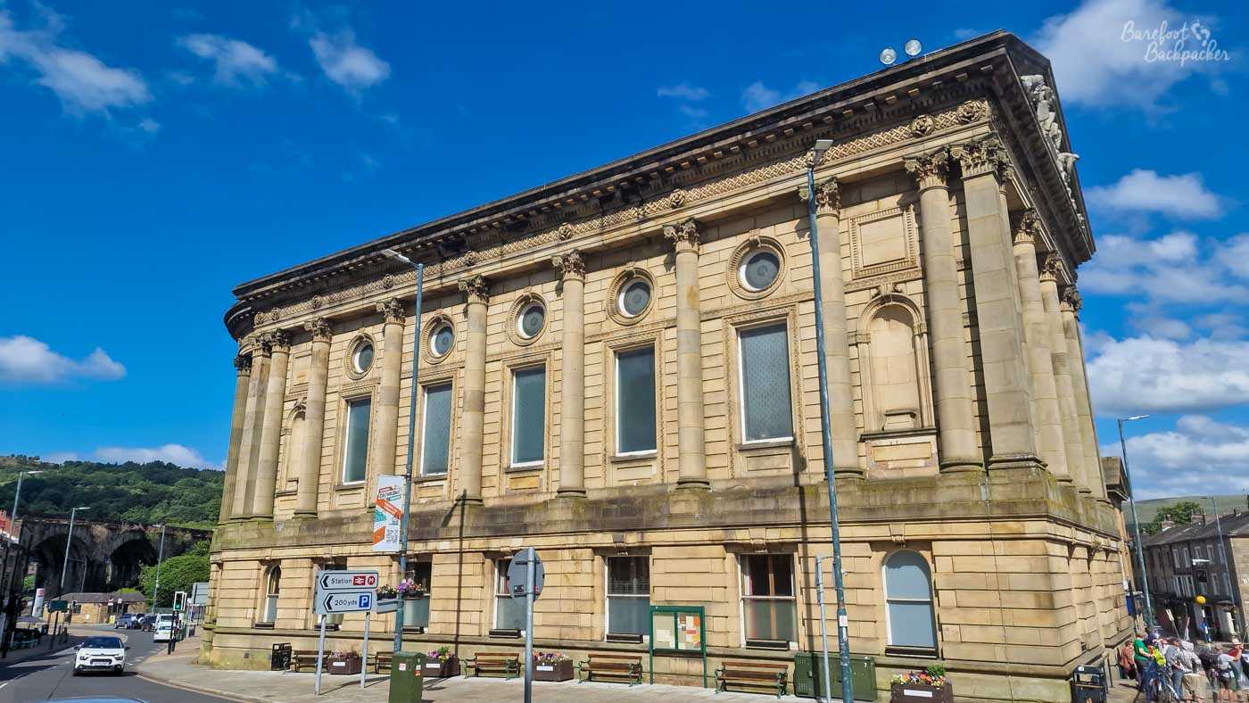 The imposing sandstone façade of Todmorden Town Hall fills the frame beneath a vivid blue sky dotted with white clouds. Designed in a grand classical style, the nineteenth-century building is lined with tall Corinthian columns, circular windows and elaborate carved stonework, giving it the appearance of a monumental civic palace. Traffic signs, benches and a handful of pedestrians at street level provide a sense of scale while hinting at its continuing role at the heart of the town. Bright sunshine emphasises the warm golden colour of the Yorkshire stone and the intricate architectural details across the façade.