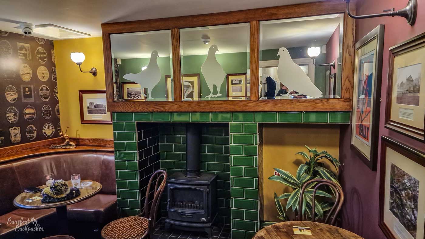 A cosy seating area centred on a wood-burning stove set into glossy green tiled surrounds, with frosted pigeon silhouettes decorating the glass partition above. Curved leather banquette seating, framed historic photographs and brewery memorabilia fill the surrounding walls, creating a comfortable corner that blends heritage features with traditional pub character.