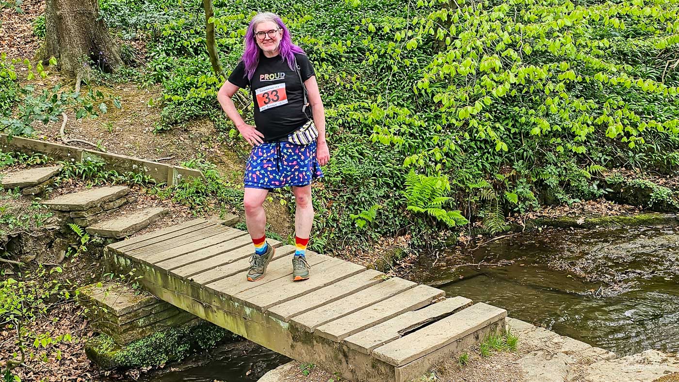 A person with purple hair stands on a wooden bridge in a forest. They are wearing a black t-shirt with the word