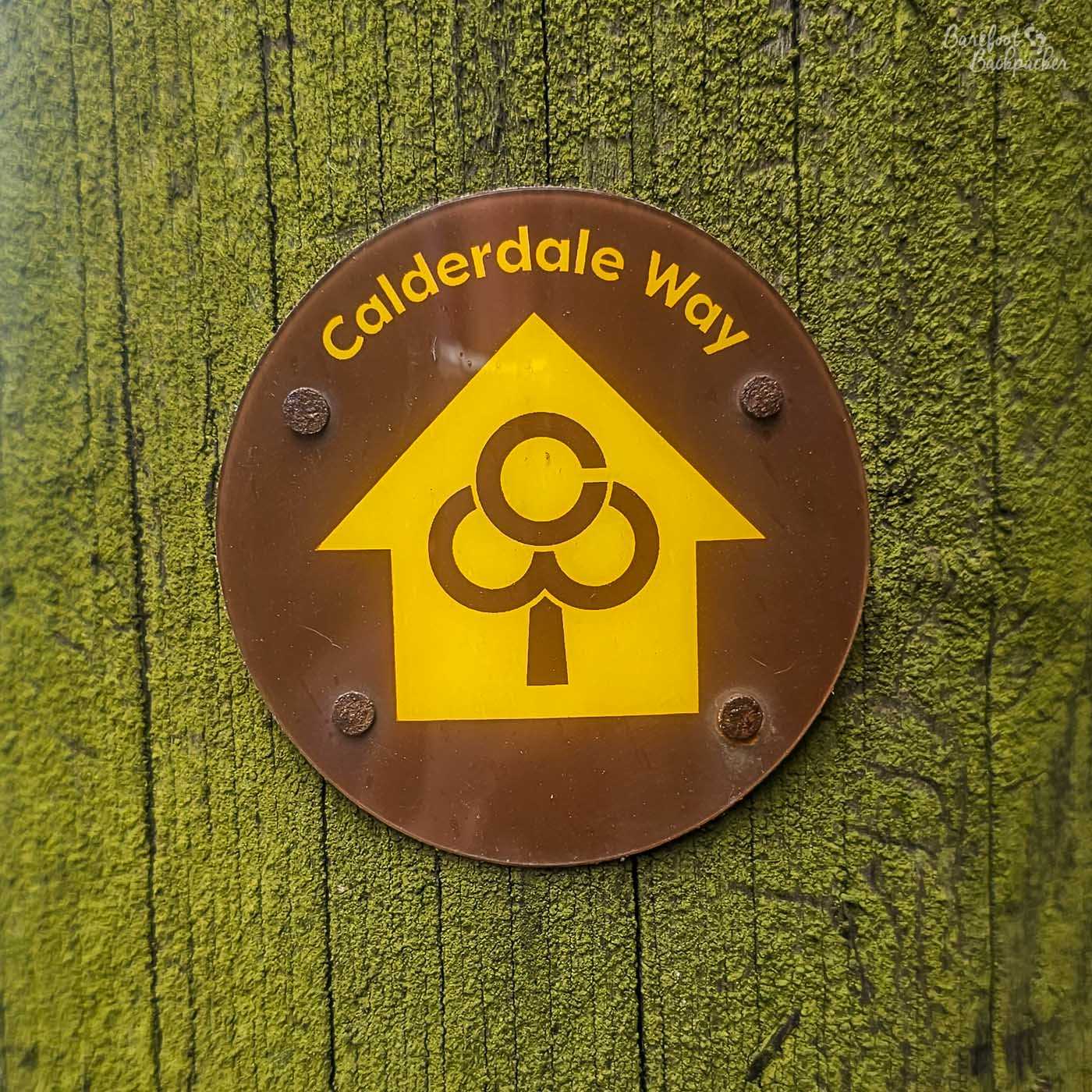 A circular brown and yellow waymarker for the Calderdale Way is fixed to a weathered wooden post coated in bright green moss. The sign features the trail's distinctive yellow arrow and stylised tree logo, with the words