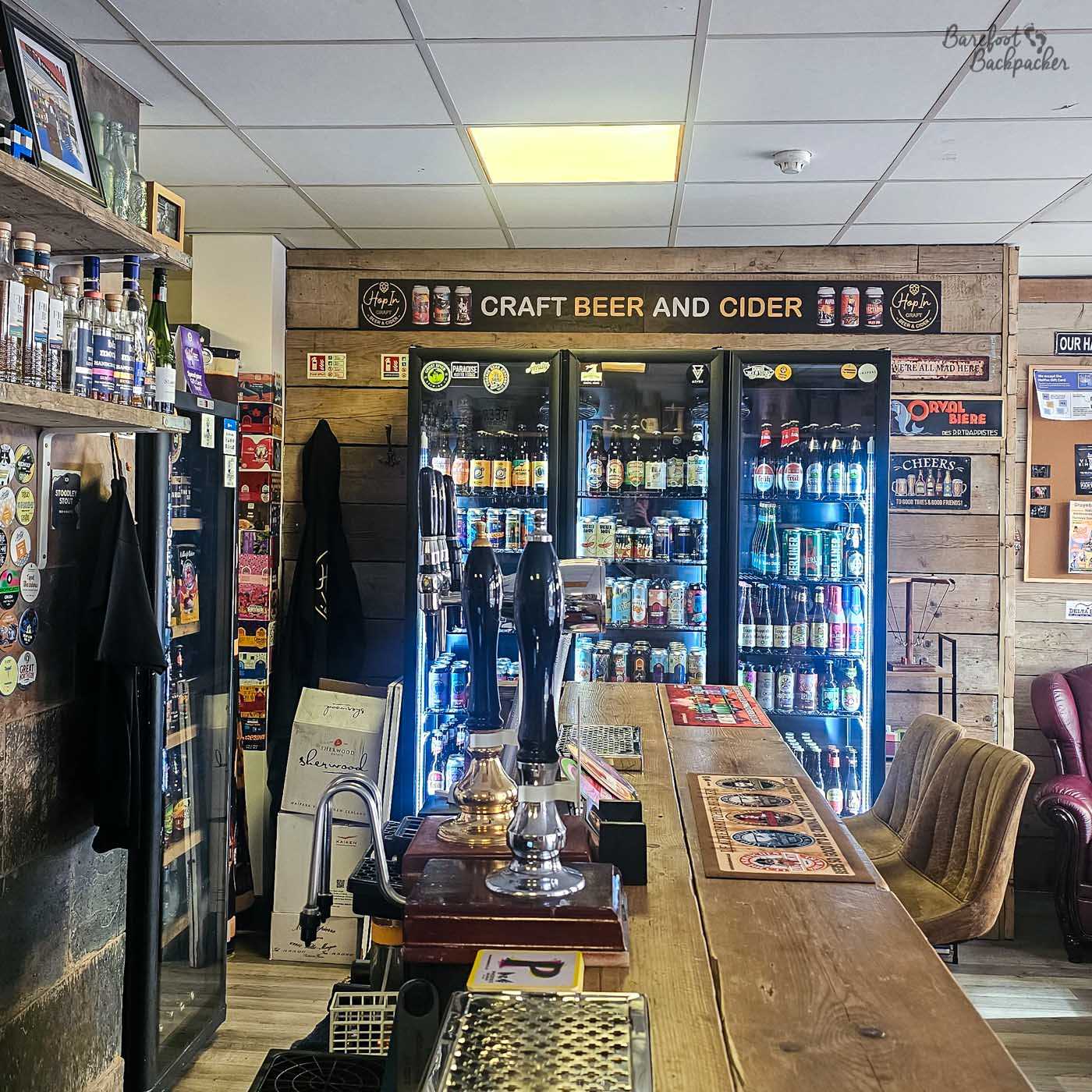The tiny interior of Hop In centres on a narrow wooden bar facing fridges filled with bottled and canned craft beers from a wide range of breweries. Hand pumps, rustic timber cladding and shelves displaying bottles and local memorabilia maximise the limited space, while a handful of chairs provide cosy seating around the edges. Every part of the micropub is dedicated to showcasing its beer selection.