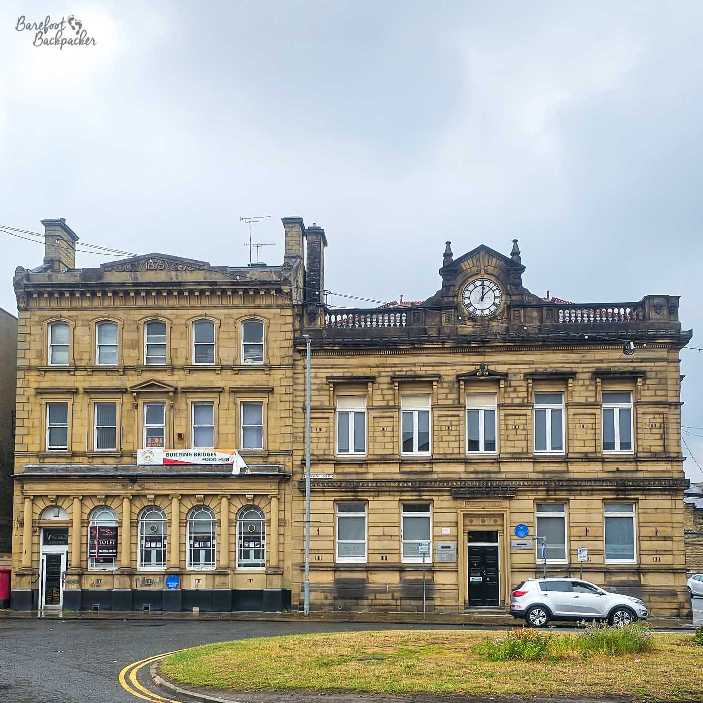 Two adjoining Victorian sandstone buildings dominate a town square, their honey-coloured façades decorated with ornate stonework, tall sash windows and classical architectural details. The building on the right is crowned by an elegant clock set into an elaborate pediment, while the neighbouring building bears the date 1875 carved into its roofline. A small patch of grass and a parked white car occupy the foreground beneath a pale grey sky, contrasting the busy road with the grandeur of the nineteenth-century architecture. The scene captures the civic confidence of former mill towns, where impressive public buildings still define the town centre long after their original purpose has changed.