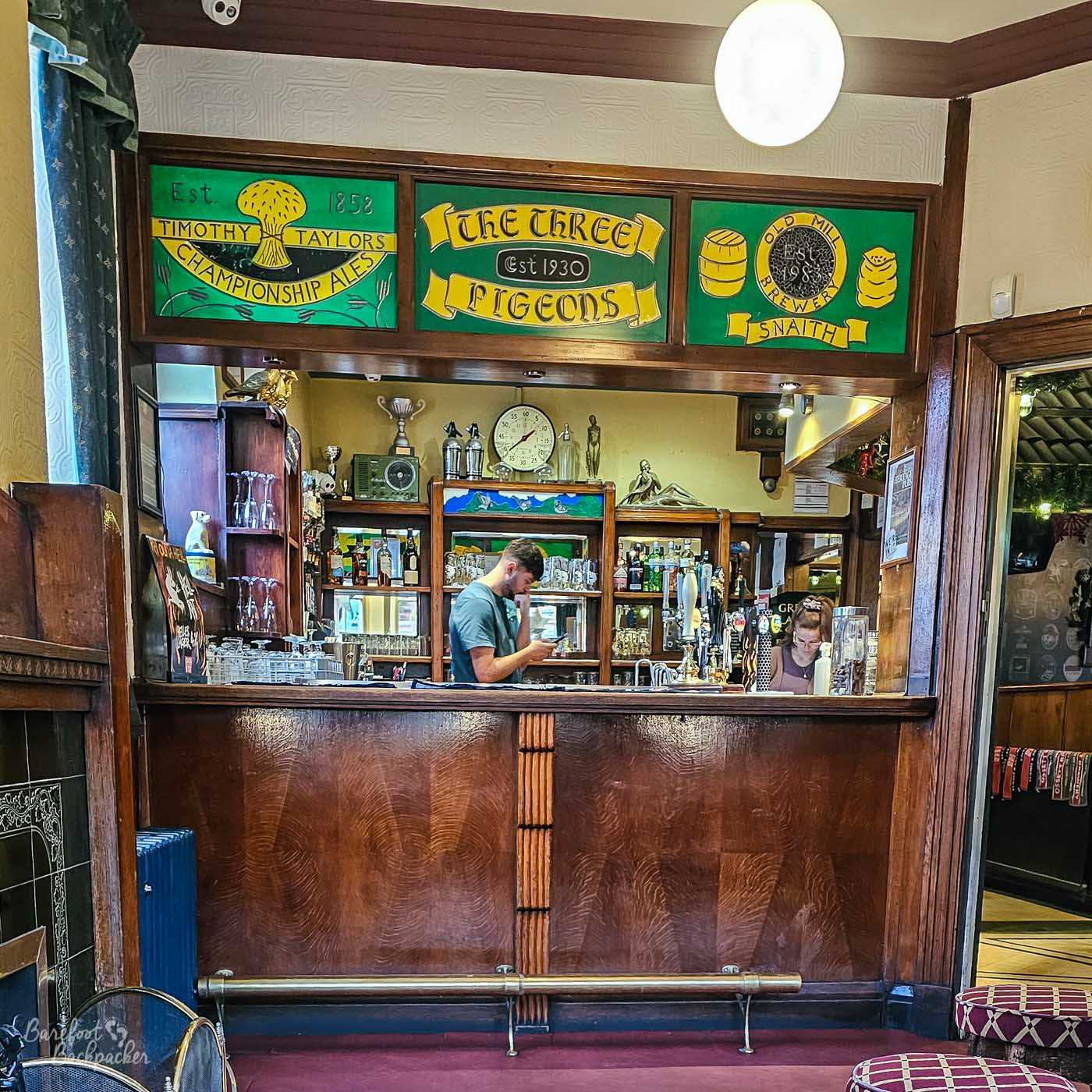 The traditional wooden bar at The Three Pigeons, framed by decorative stained-glass panels celebrating the pub and local breweries. Behind the counter, shelves display bottles and glassware while two members of staff prepare drinks beneath vintage clocks, trophies and ornaments. Dark polished wood and warm colours give the room a classic community pub feel.