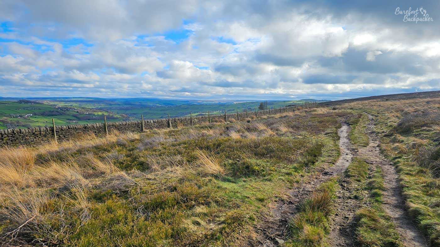 A rough twin-track path curves gently across open moorland, bordered by heather, coarse grasses and a long dry stone wall that follows the contours of the hillside. Beyond the wall, green valleys and distant hills unfold beneath a sky filled with towering white and grey clouds, where patches of blue break through to illuminate parts of the landscape. The contrast between the rugged foreground and the softer farmland below highlights the transition between wild uplands and cultivated countryside. The winding track invites the viewer onwards, suggesting the countless routes that cross the Pennine moors.