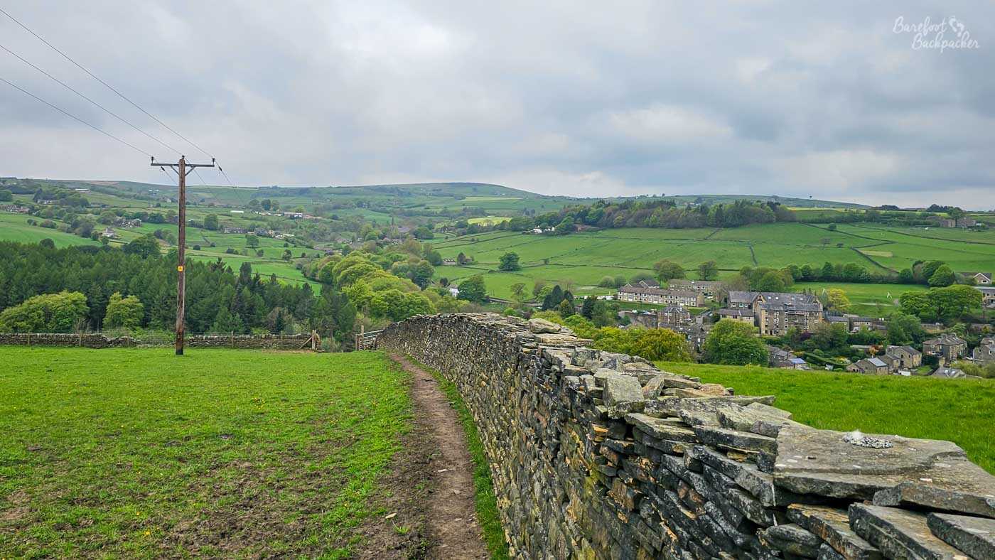 A narrow footpath follows the line of a beautifully built dry stone wall across bright green pasture overlooking a village nestled in the valley below. Clusters of trees surround stone houses and fields divided into neat patchwork patterns, while rolling hills continue into the distance beneath a blanket of grey cloud. A solitary wooden telegraph pole rises beside the path, adding a simple vertical landmark against the expansive countryside. The elevated viewpoint showcases the distinctive Pennine landscape where settlements occupy the valley floor and farmland climbs steadily up the surrounding hillsides.