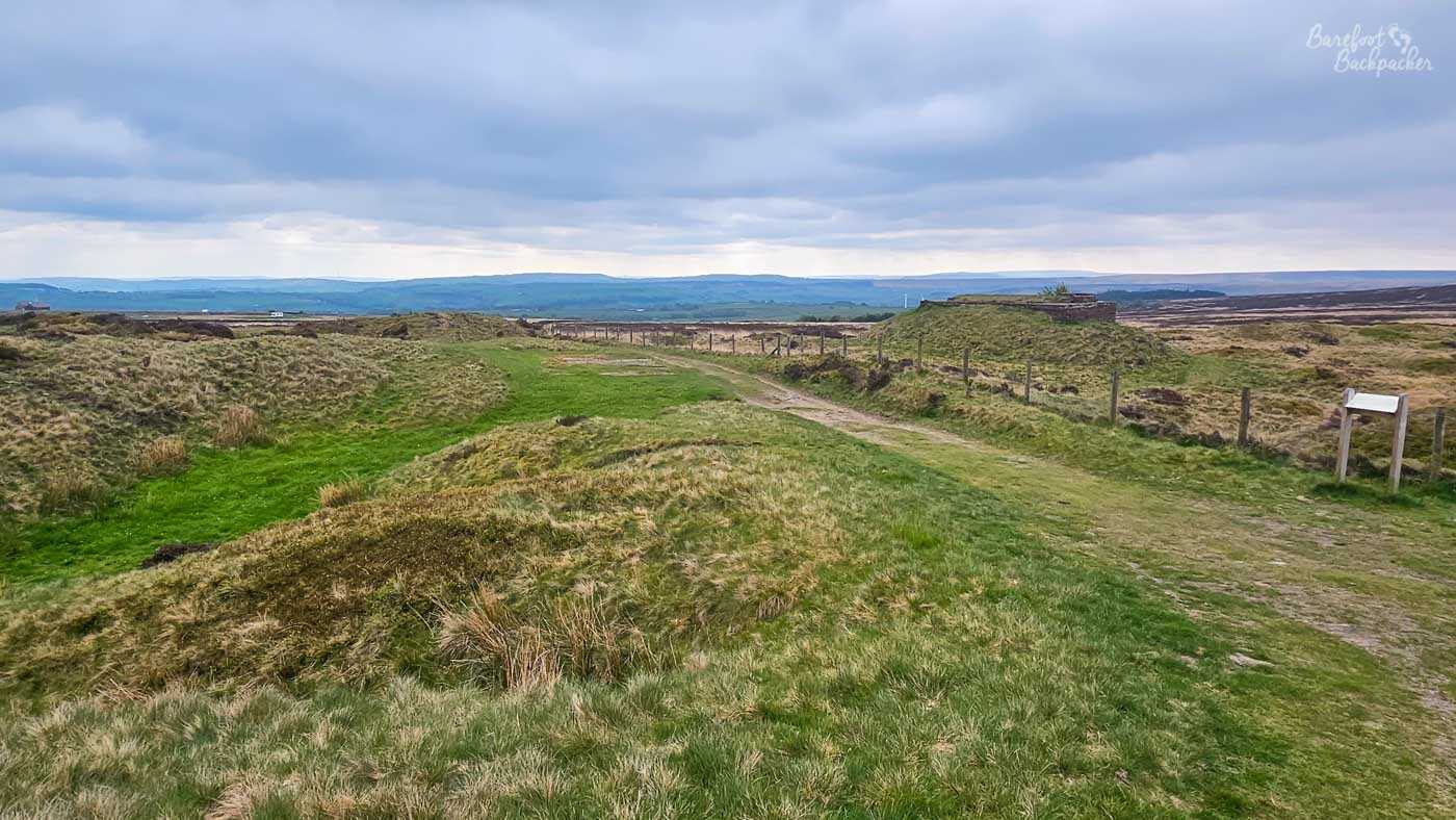 A grassy track winds across open moorland where low, uneven mounds ripple across the foreground before the land falls away towards distant hills. A small stone structure sits on a raised bank to one side while information boards and fence lines hint at the historic significance of the landscape, despite its quiet appearance today. Heavy clouds gather overhead, casting soft shadows across the rough grass and heather, while far beyond the moors the countryside fades into layers of blue-grey hills. The broad, open setting conveys both the remoteness of the uplands and the traces of human history still preserved within them.