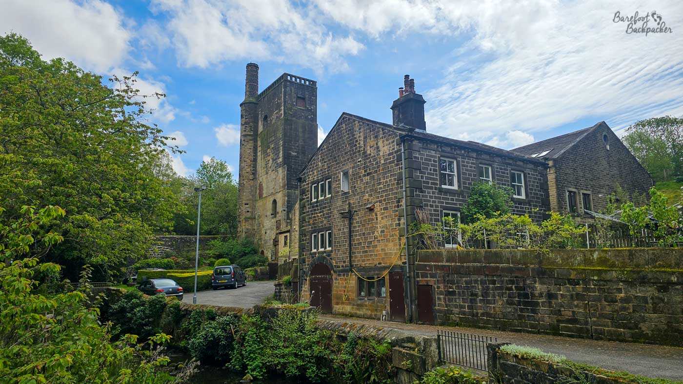 A tall stone mill chimney rises above a cluster of dark gritstone buildings beside a narrow stream, standing as a reminder of the area's industrial past. Mature trees frame the scene with fresh spring foliage while patches of moss and weathering soften the old masonry, contrasting with the neatly maintained houses now occupying part of the former mill complex. A bright blue sky streaked with delicate clouds brings warmth to the otherwise rugged architecture, highlighting the blend of industry and countryside that characterises many Pennine valleys. The scene captures how former textile mills have become woven into the everyday landscape rather than standing apart from it.