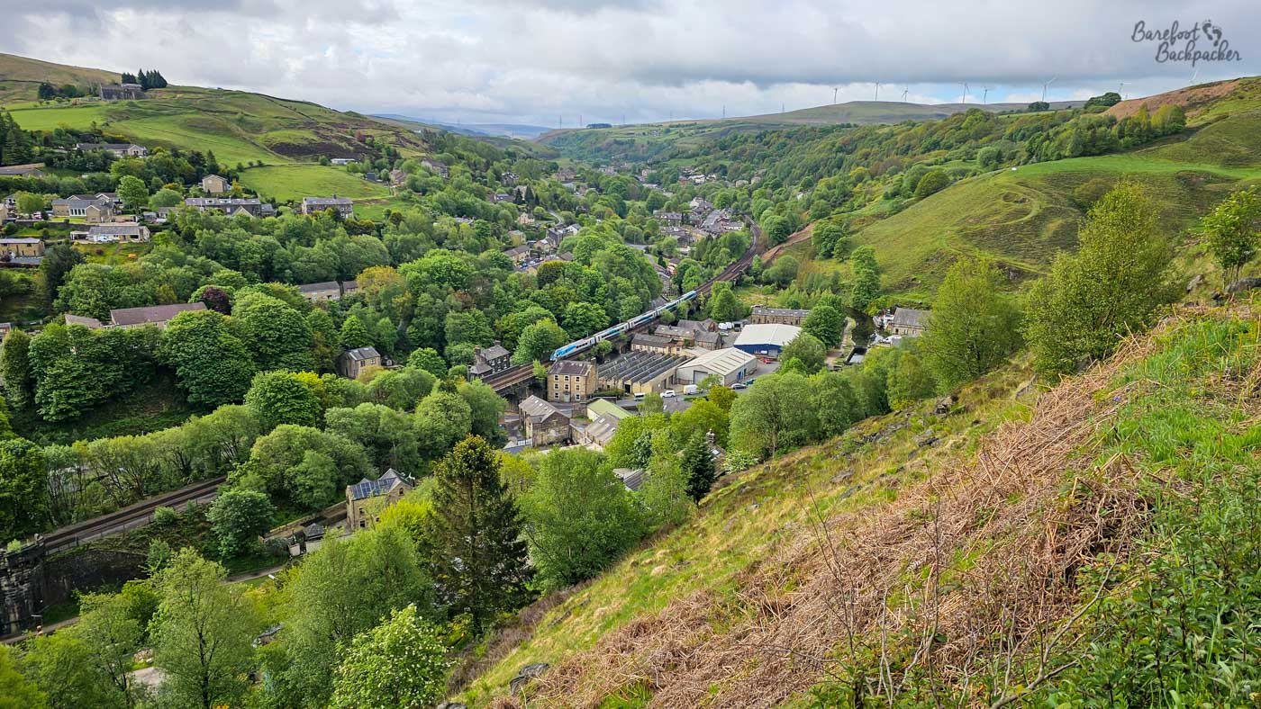 Looking down from a steep grassy hillside, a railway line threads through a densely wooded valley where clusters of stone buildings gather around the tracks. A blue passenger train crosses the centre of the scene, dwarfed by the surrounding hills, while roads, houses and mills nestle among the trees below. Beyond the settlement, the valley narrows towards distant moorland where a line of wind turbines stands along the skyline beneath heavy clouds. The elevated viewpoint clearly shows how the transport routes, settlements and woodland all follow the contours of the valley through the Pennines.
