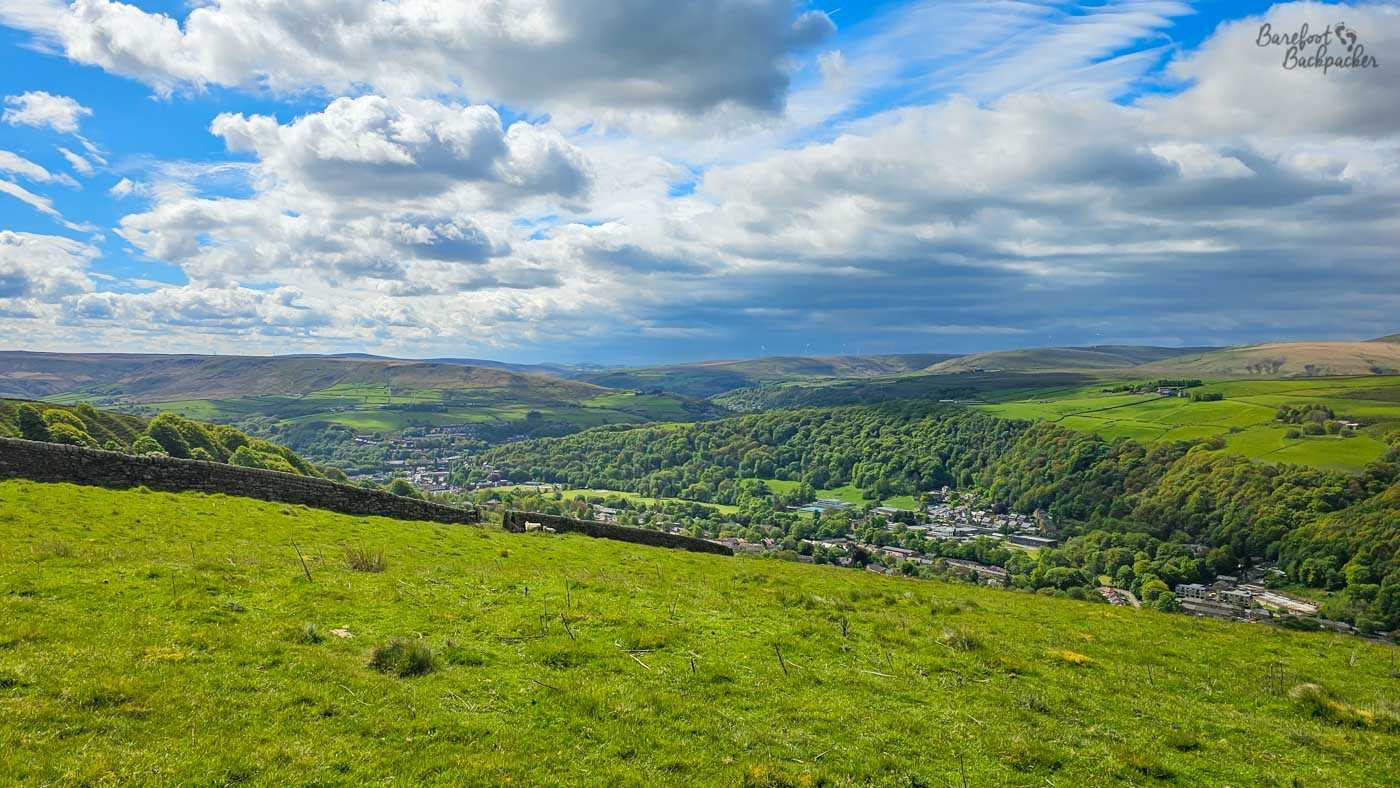 A sweeping panoramic view stretches across a broad Yorkshire valley filled with patchwork green fields, wooded hillsides and a town nestled below. Dry stone walls divide the foreground pasture before the land drops away into dense woodland surrounding the valley floor, while rolling moors extend across the horizon under a sky packed with towering white and grey clouds. Sunlight breaks through in places, illuminating sections of the landscape while leaving others in shadow, giving the view a dramatic sense of depth and constantly changing weather. The scene captures both the openness of the uplands and the villages tucked into the sheltered valleys below.