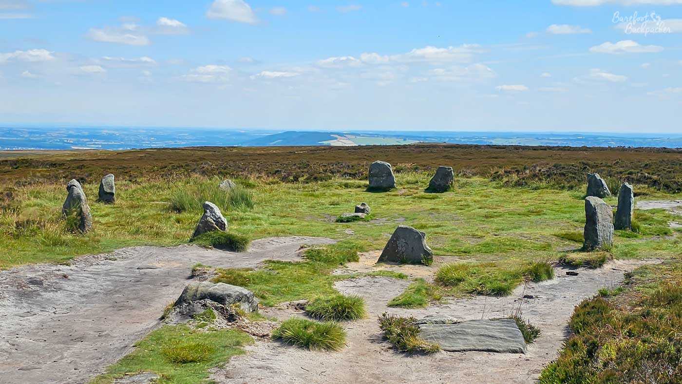 A wide, panoramic shot captures a cluster of large, ancient-looking stones standing upright in a grassy, moorland landscape. The stones, varying in size and shape, are scattered across the terrain, with some appearing to form a rough circle. The ground around the stones is a mix of dry, sandy paths and patches of vibrant green grass, with some areas of heather or gorse adding a touch of brown and purple to the vegetation. In the background, the land slopes gently downwards, leading to a hazy, distant landscape of fields and possibly a town or city, all under a bright blue sky dotted with fluffy white clouds.