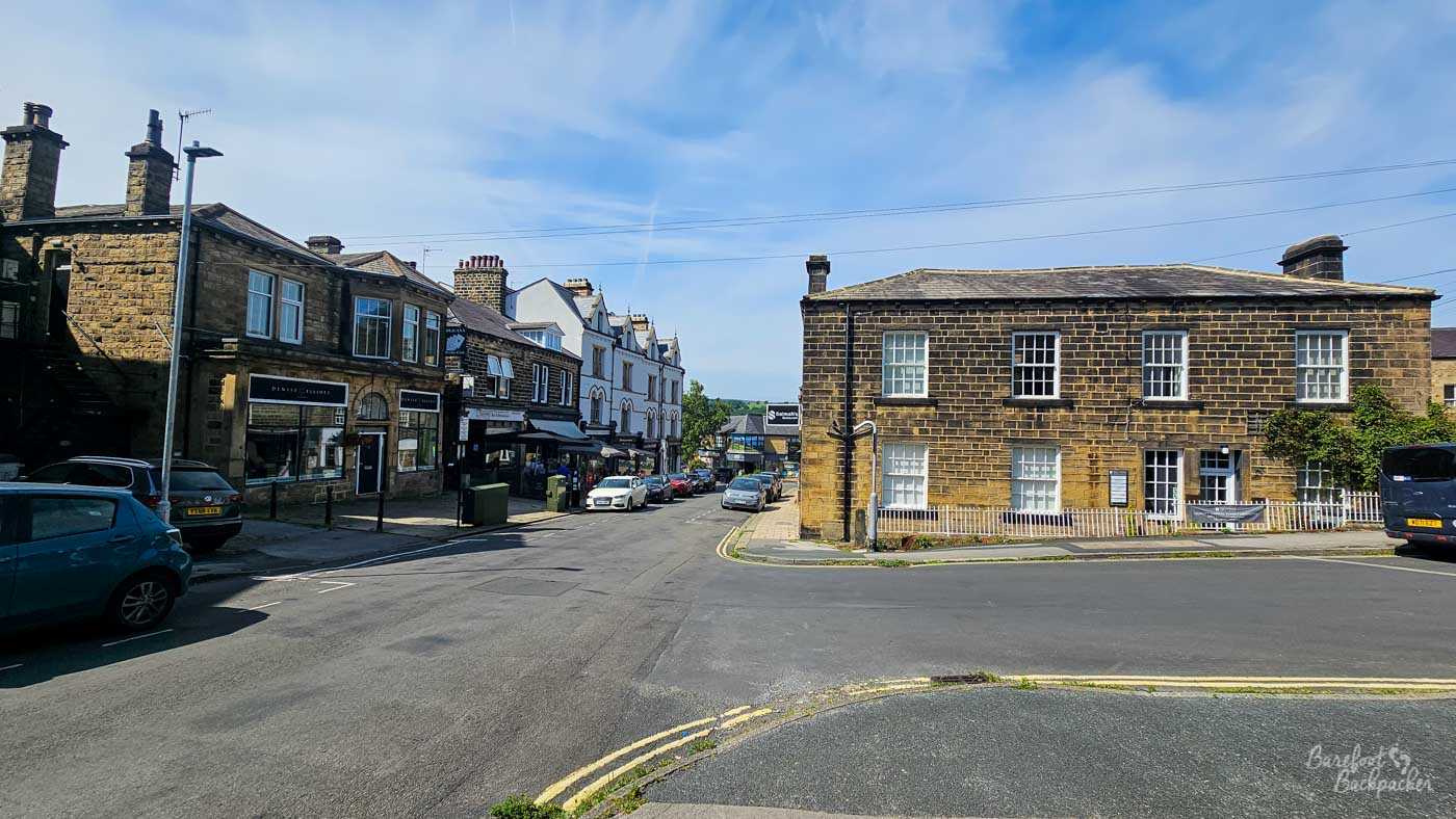 A warm, sunlit view captures a picturesque street scene in a charming village, likely in the UK, given the architectural style. The image is dominated by traditional stone buildings with multi-paned windows and classic chimneys, a testament to enduring craftsmanship. On the right, a prominent two-story stone building with white-framed windows stands out. A white picket fence lines its front, adding a touch of quaintness, and a dark bus is parked to its right. To the left, a row of similar stone buildings houses various shops and businesses. One building features a dark awning over its storefront, and several cars are parked along the street, including a teal-colored car in the foreground and a dark SUV behind it. People are visible dining or browsing at the establishments, suggesting a lively atmosphere. The road, paved with asphalt, is marked with yellow double lines, indicating a junction. The sky above is a bright blue, adorned with wispy white clouds, hinting at a pleasant summer day. The overall impression is one of a well-preserved, vibrant village community basking in the glow of a beautiful day.