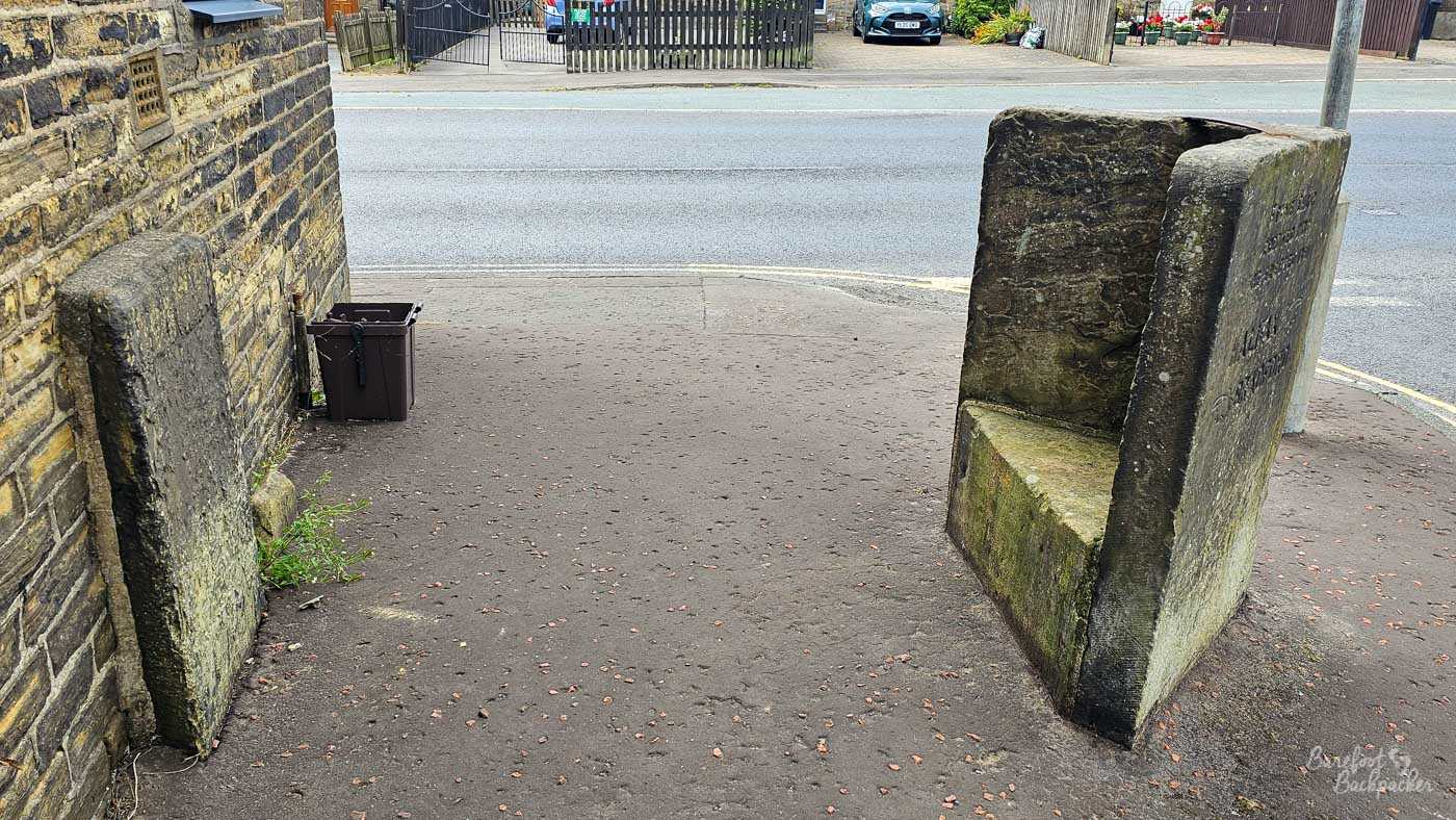A low-angle, slightly wide shot shows a weathered stone structure on a sidewalk. The structure, possibly a historic marker or bench, has a rough, moss-covered surface and is made of large, rough-hewn stone blocks. It is positioned on the right side of the frame, with a stone pillar leaning against a brick wall on the left. A dark trash bin is visible between the pillar and the wall. The sidewalk is made of asphalt with small reddish stones embedded in it. Behind the sidewalk, a gray asphalt road runs horizontally across the image. On the other side of the road, a dark wooden fence encloses a property with a modern dark blue car parked in front of it. The sky is overcast and appears to be a soft gray.