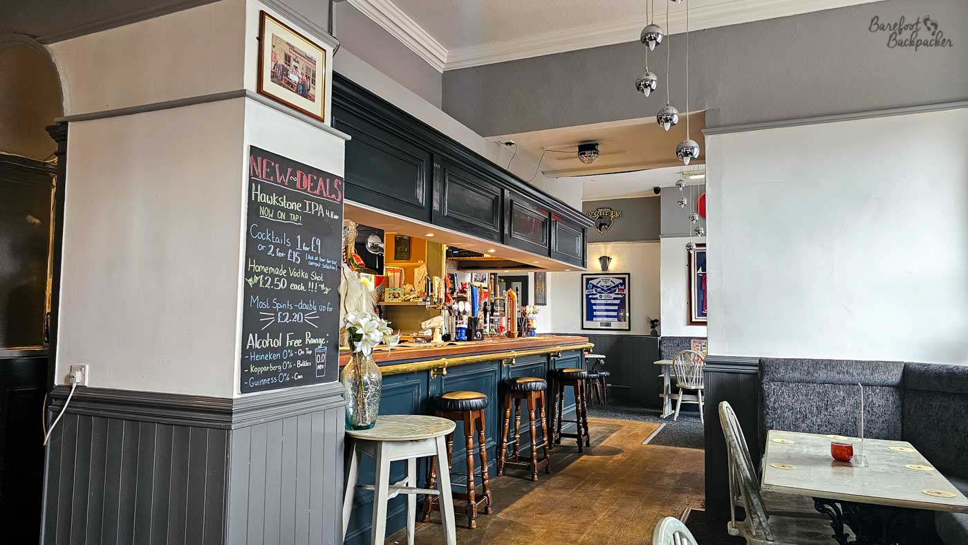 A traditional pub interior with a dark wood bar occupying one side of the room, hand pumps displaying cask ales and comfortable seating arranged beside large front windows. Grey upholstered benches, framed local memorabilia and polished wooden flooring create a relaxed atmosphere, while daylight streams in from the road outside.