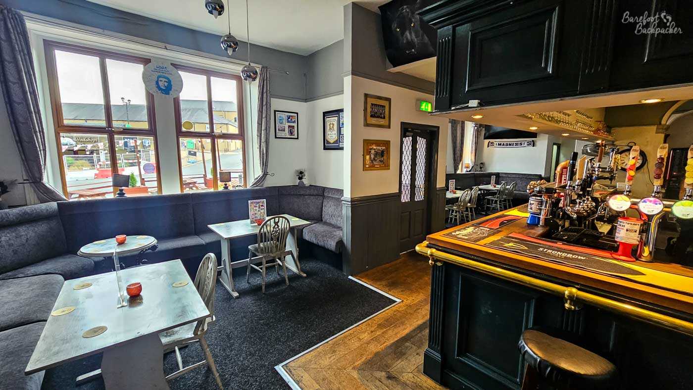 A wider view across the main lounge shows the traditional bar framed by dark wood panelling, with stools lining the counter and comfortable seating spread throughout the room. Chalkboards advertise drinks offers while framed sports memorabilia and photographs decorate the walls. Neutral colours, soft lighting and spacious seating create the feel of a welcoming local community pub.
