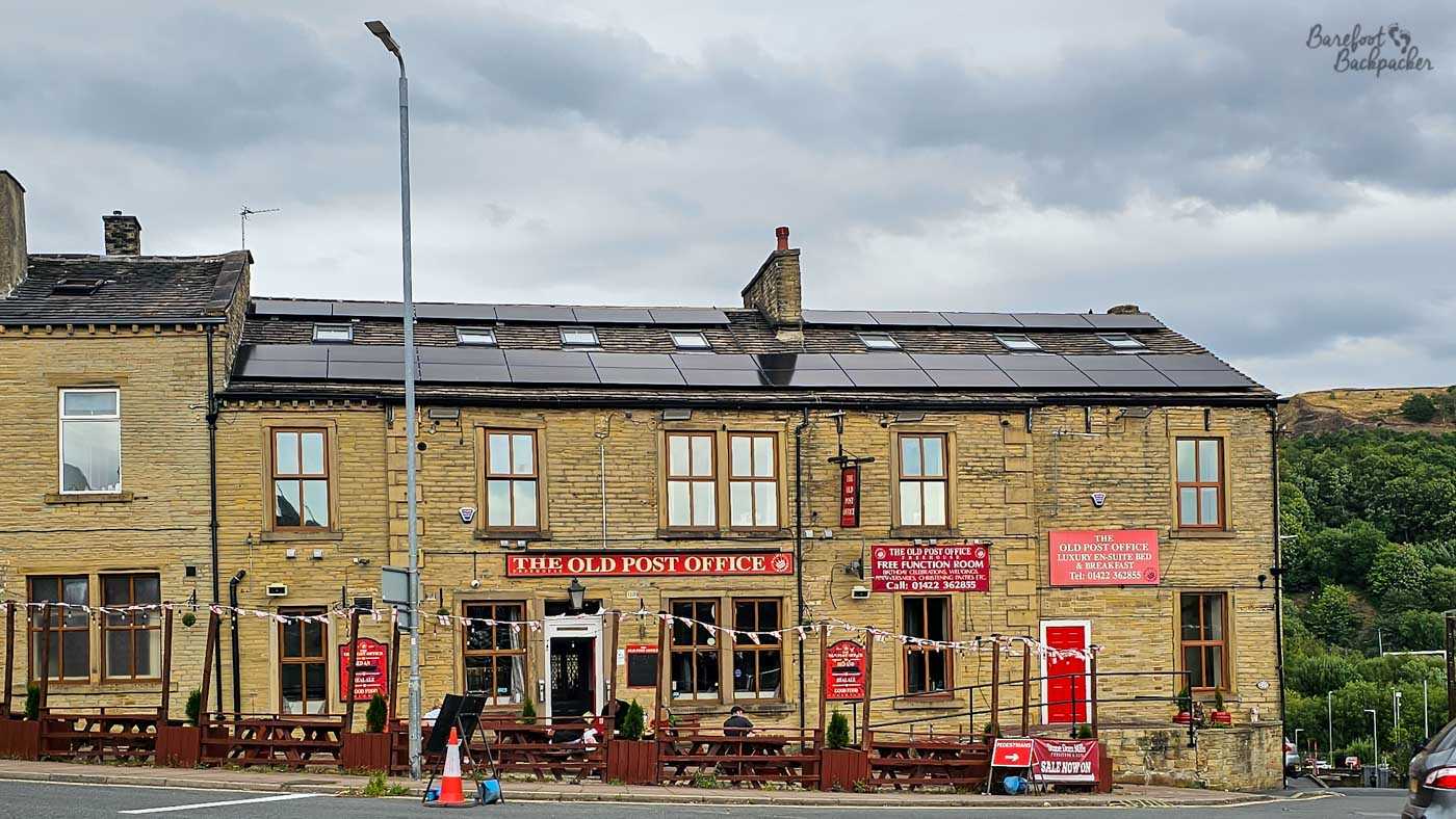 The Old Post Office occupies a substantial stone building overlooking the roadside, with traditional Yorkshire stone walls, red signage and rows of outdoor picnic benches. Solar panels line the roof while wooded hills rise behind the pub, placing it on the edge of the Calder Valley landscape. The building also offers accommodation alongside its pub facilities.