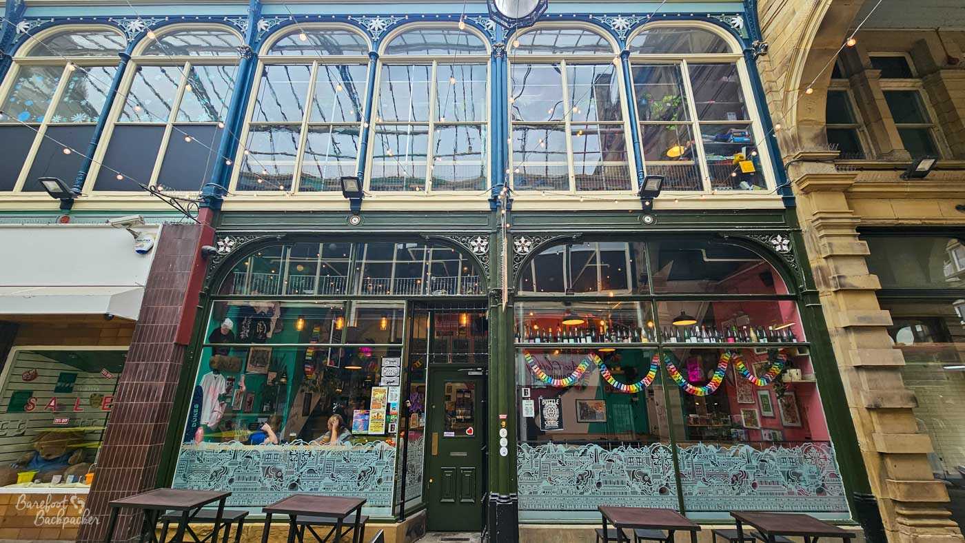 The striking frontage of Dukes occupies a tall Victorian arcade building with ornate ironwork, huge glazed windows and decorative upper-storey arches. Rainbow paper decorations and colourful seating are visible through the expansive windows, while small outdoor tables sit beneath the covered arcade. The impressive historic architecture contrasts with the venue's bright contemporary interior.