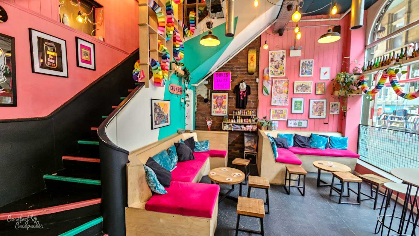A bright seating area with vivid pink walls, turquoise accents and colourful artwork covering almost every available surface. Built-in benches with bold pink cushions line the walls beneath strings of rainbow paper chains, while a staircase curves upwards towards an upper level. Large front windows flood the room with natural light, making the venue feel cheerful and creative.