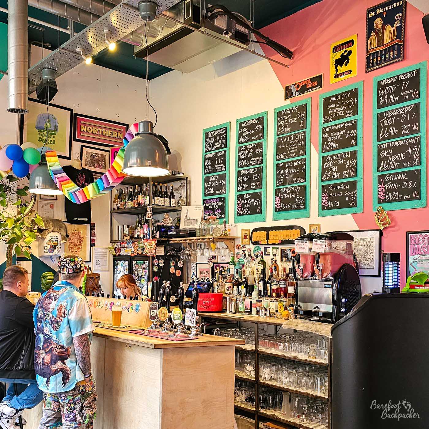 The lively main bar at Dukes is filled with colourful artwork, chalkboards listing an extensive rotating beer selection and shelves stocked with spirits and bottles. Bright pink and turquoise walls, industrial lighting and rainbow paper decorations give the venue a playful, welcoming atmosphere, while customers chat at the counter beneath large handwritten beer boards. The décor reflects the bar's independent, alternative character.