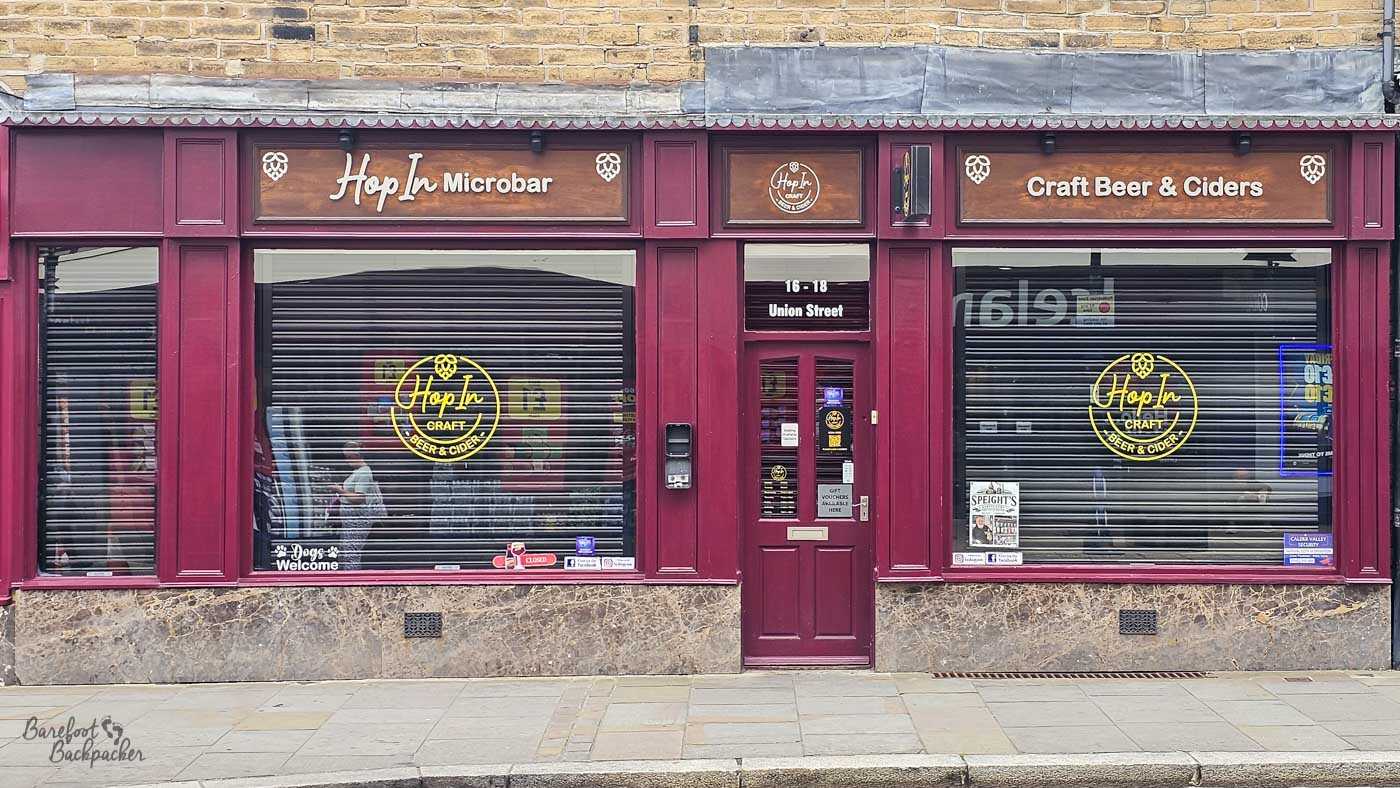 The compact frontage of Hop In Micropub, with deep burgundy painted woodwork framing large front windows displaying the pub's craft beer and cider branding. The simple, symmetrical exterior reflects the intimate size of the venue, while signs advertise craft beers, ciders and a dog-friendly welcome. From the street it resembles a traditional shop more than a conventional pub.