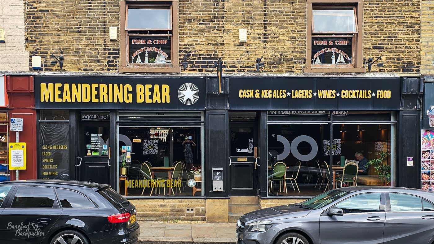 The black-painted frontage of Meandering Bear stands beneath the upper storeys of an older stone building, with bold yellow lettering advertising cask ales, lagers, wines, cocktails and food. Large windows reveal the contemporary interior, while parked cars line the street outside and a customer sits quietly inside by the window. The exterior combines modern branding with the character of the historic town centre.