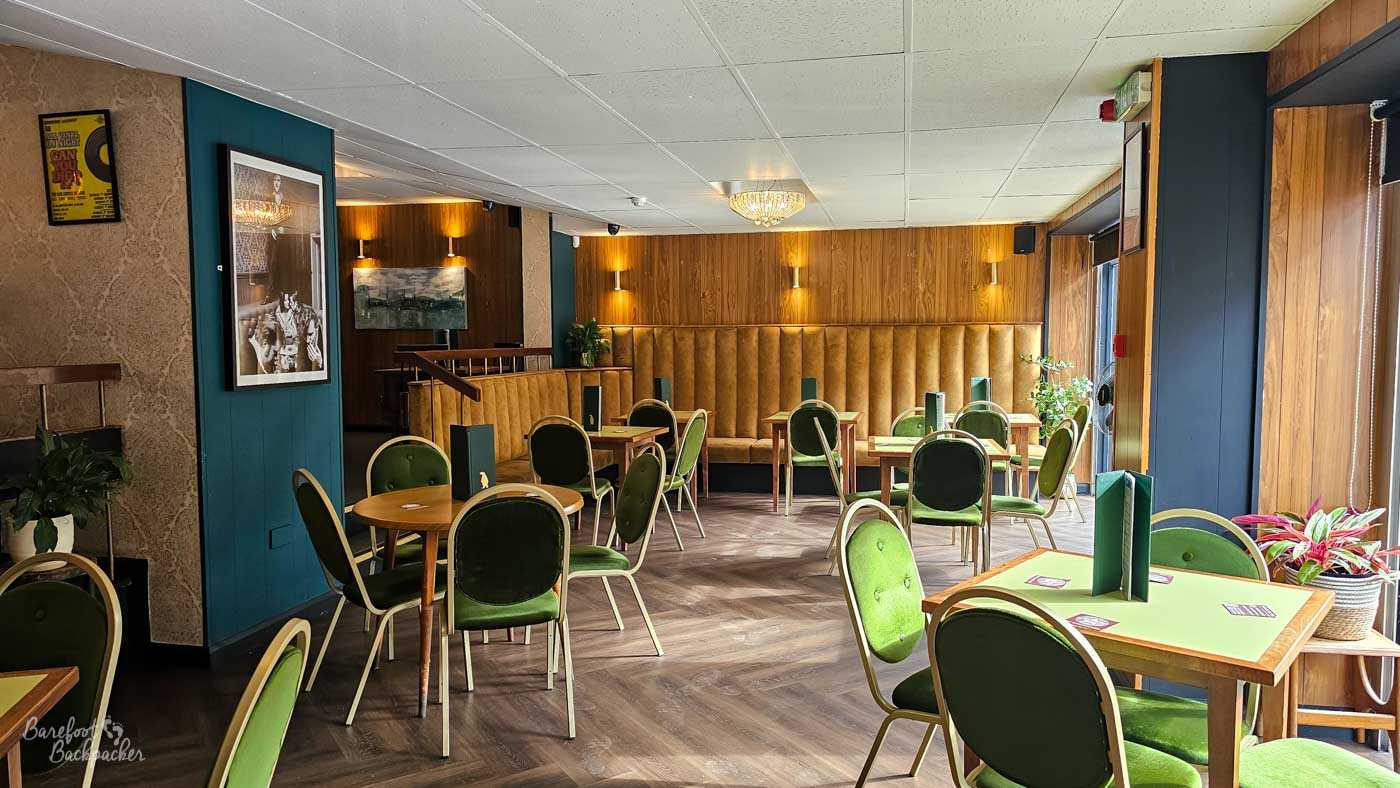 A spacious seating area with green upholstered chairs, wooden tables and long high-backed banquette seating running along one wall. Warm timber panelling, subtle lighting and contemporary décor create a relaxed atmosphere, with generous spacing between tables giving the venue an open, comfortable feel. The modern design contrasts with the traditional stone building that houses the pub.