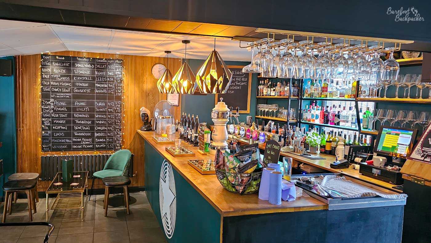 The modern bar at Meandering Bear features a polished wooden counter beneath striking geometric pendant lights and rows of hanging wine glasses. A large blackboard lists the current beers on tap alongside breweries, strengths and prices, while shelves behind the bar display spirits, wines and craft beers. Clean contemporary styling is balanced by warm wood finishes and soft lighting.