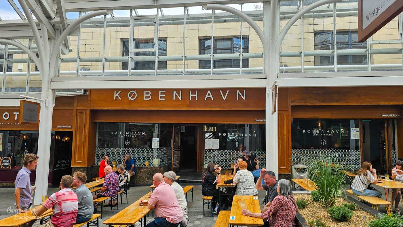Outdoor seating in the covered Westgate Arcade outside København, where long wooden benches are filled with people enjoying drinks and conversation. The pub's broad frontage combines warm timber cladding with large windows displaying the København branding, while planters and the glass-roofed arcade create a sheltered courtyard atmosphere in the heart of the city.