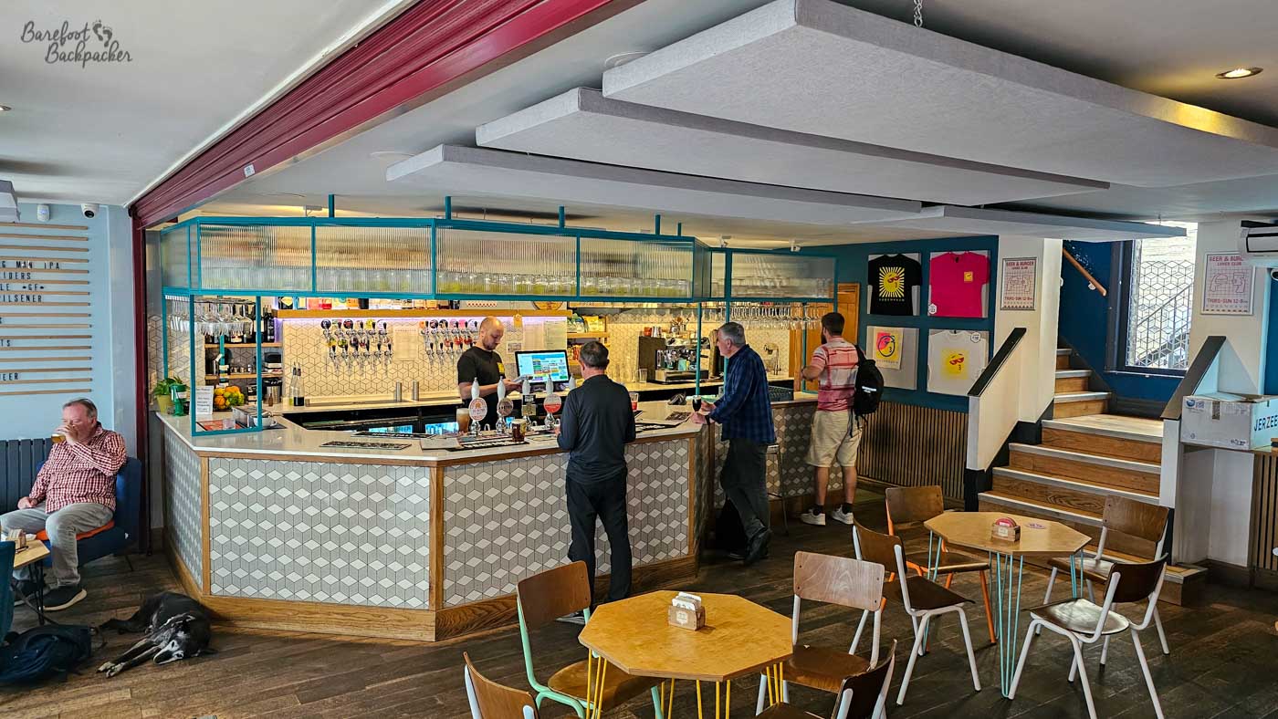 The central bar at København, finished with geometric patterned tiles and pale wood beneath rows of hanging glasses. Several customers chat with staff while ordering drinks, and modern seating spreads across the open-plan room, including a relaxed seating area where a black dog sleeps peacefully on the floor. Clean lines, bright colours and contemporary design create a relaxed craft beer atmosphere.