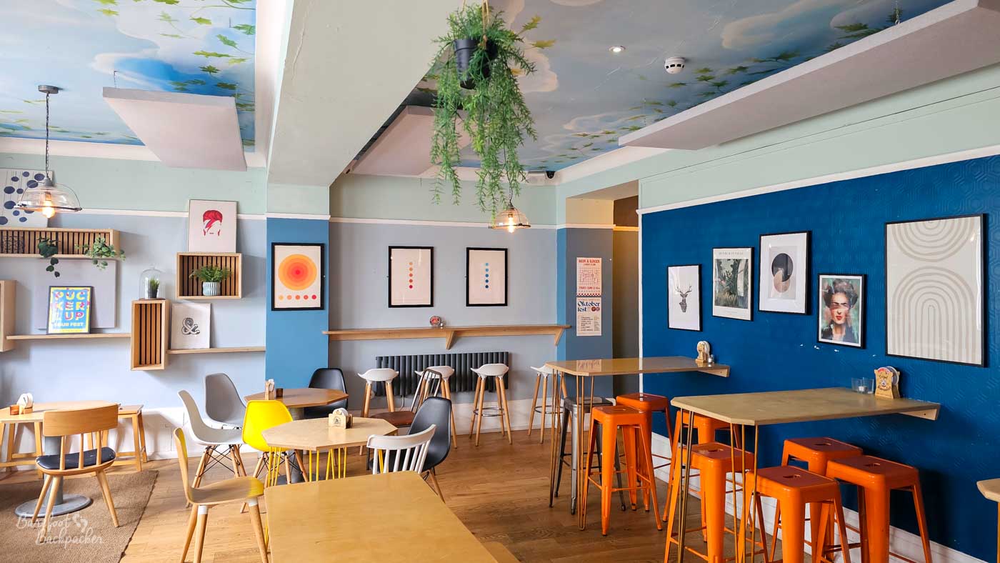 A bright, contemporary seating area with pale wooden tables, colourful chairs and deep blue feature walls decorated with modern prints. Hanging plants, shelves with small ornaments and a painted ceiling featuring blue sky and clouds give the room a light, Scandinavian-inspired feel. The spacious layout contrasts with the traditional appearance of many neighbouring pubs.