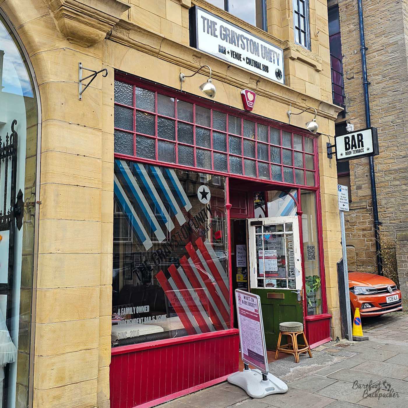 The entrance to Grayston Unity, housed within a sandstone building with deep red painted woodwork framing large front windows. A sign above describes it as a bar, venue and cultural hub, while posters and a pavement board advertise upcoming events beside the doorway. The distinctive frontage stands out among the surrounding city-centre buildings.