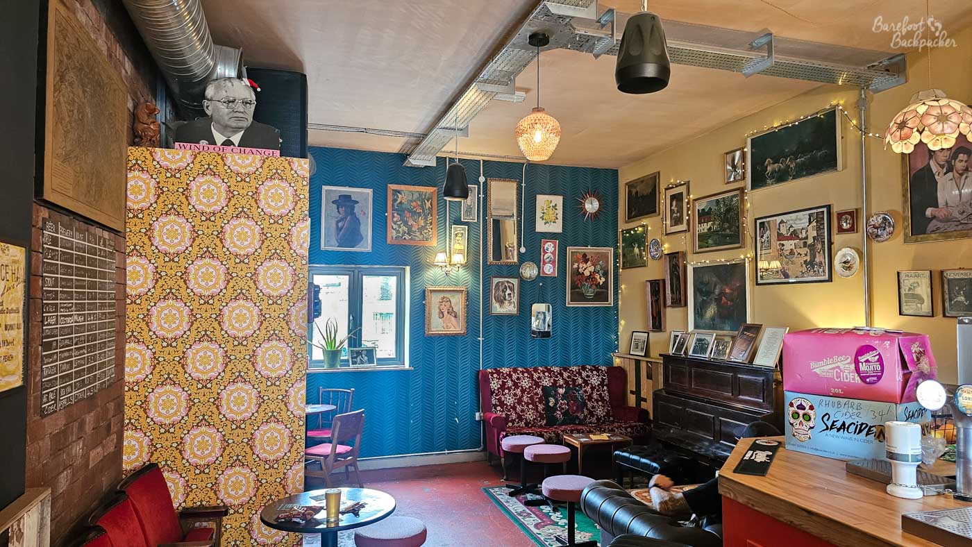 A comfortable lounge area filled with mismatched furniture, framed artwork and vintage decorations covering almost every wall. Patterned wallpaper, an upright piano, small café tables and strings of fairy lights create an informal, creative atmosphere, while portraits, mirrors and curiosities reinforce the venue's bohemian style. The room feels more like an independent arts space than a traditional pub.