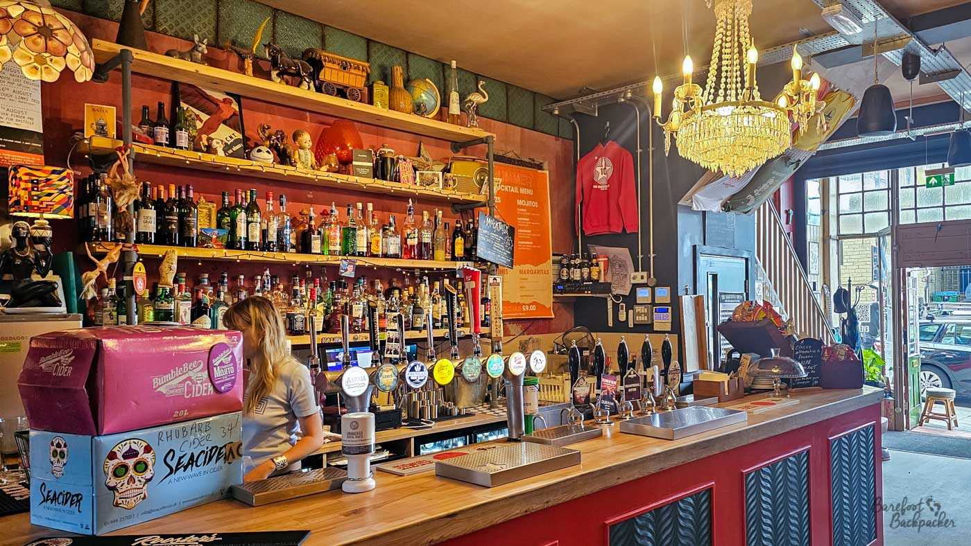 The main bar at Grayston Unity, where a long wooden counter is lined with hand pumps and backed by shelves packed with spirits, craft beers and eclectic ornaments. A crystal chandelier hangs unexpectedly above the industrial-style ceiling, while colourful decorations, vintage curiosities and memorabilia give the venue a deliberately quirky, artistic character. Daylight streams in through the open entrance, connecting the lively bar with the street outside.