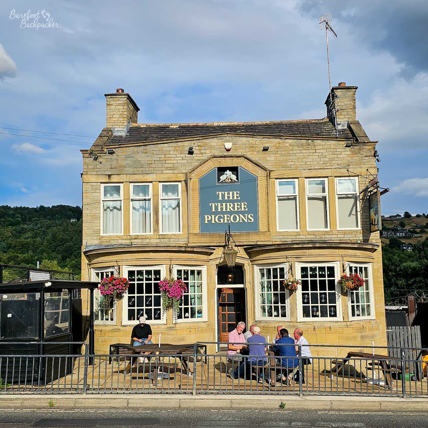 The stone frontage of The Three Pigeons stands beside the roadside beneath a blue sky, its name displayed prominently above the entrance. Hanging baskets overflowing with pink flowers brighten the pale Yorkshire stone, while several regulars sit chatting around picnic benches on the terrace in the afternoon sunshine. Low hills rise behind the pub, placing it on the edge of the surrounding valley.
