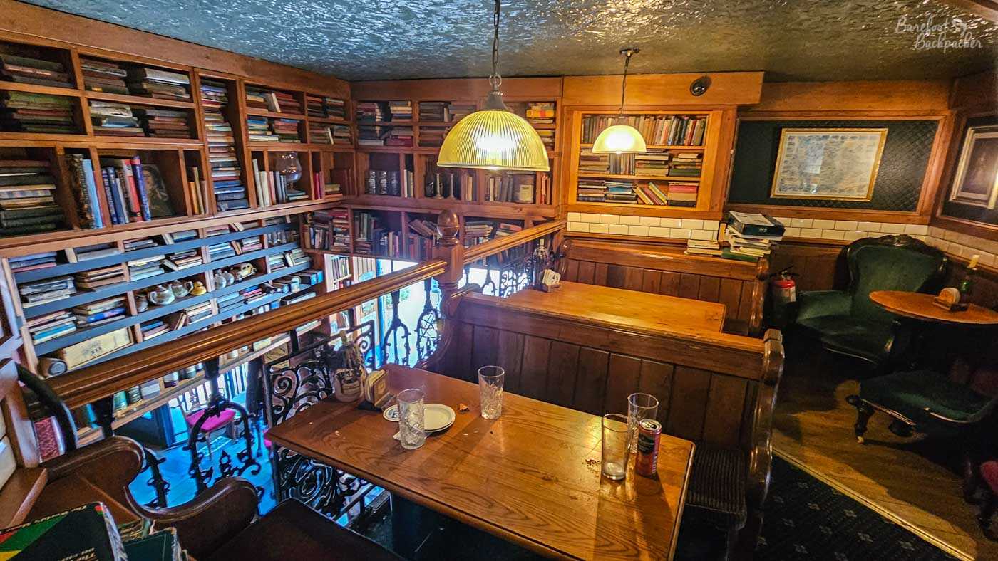 An upstairs seating area overlooking the lower floor, surrounded by wooden shelving filled with hundreds of old books. Dark wood panelling, leather armchairs and hanging pendant lights create a quiet reading-room atmosphere, with ornate iron railings opening onto the space below. Empty tables and scattered glasses suggest a relaxed afternoon between busy periods.
