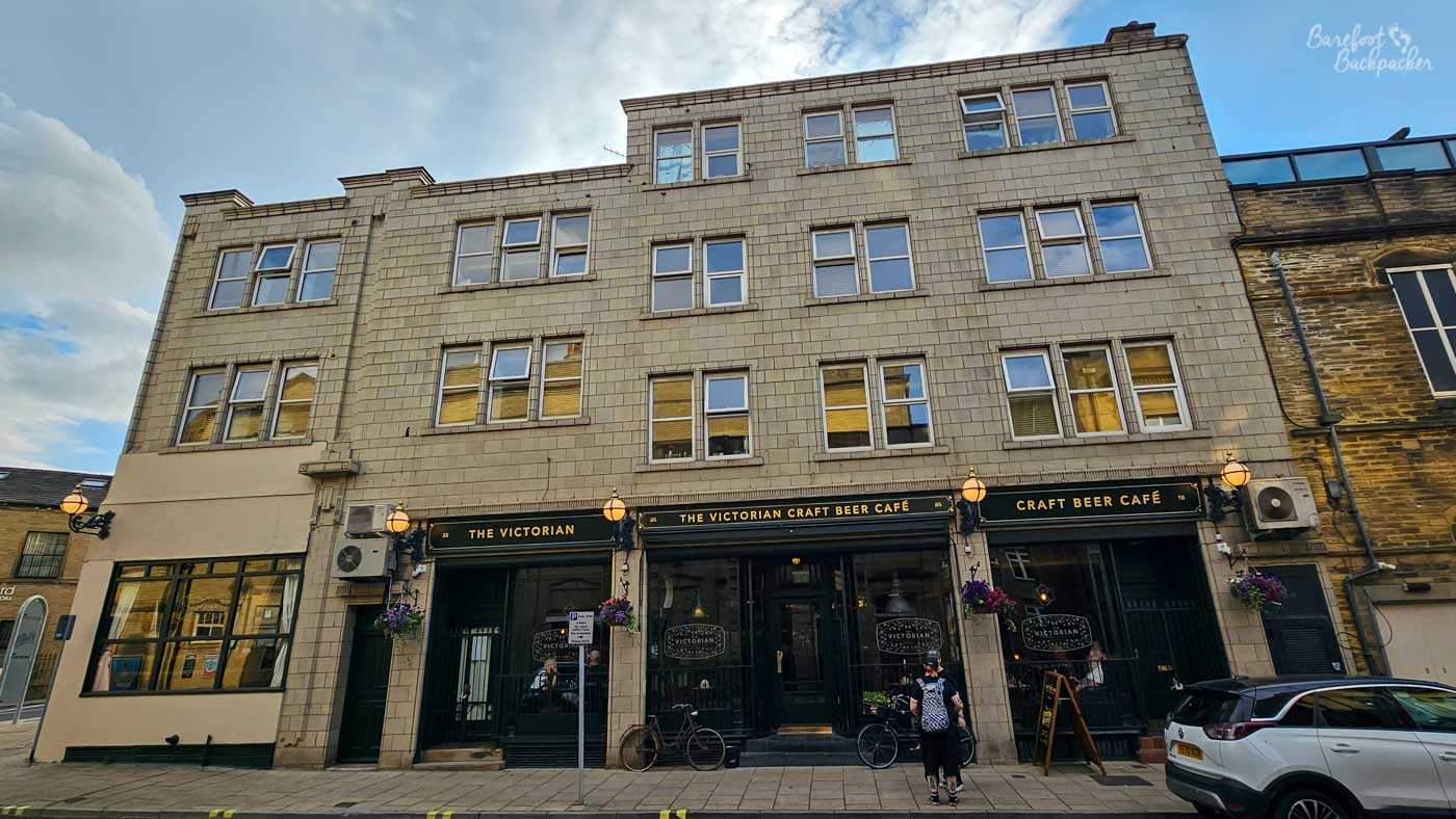 A wide street-level view of the Victorian Craft Beer Café, occupying the ground floor of a tall, pale stone building with several storeys of offices or apartments above. Dark green frontage, hanging flower baskets and traditional lamps frame the entrance, while bicycles lean against the windows and a single visitor stands outside beneath the sign. The broad façade contrasts with the intimate pub below, emphasising how easily this specialist beer bar could be overlooked within the larger building.