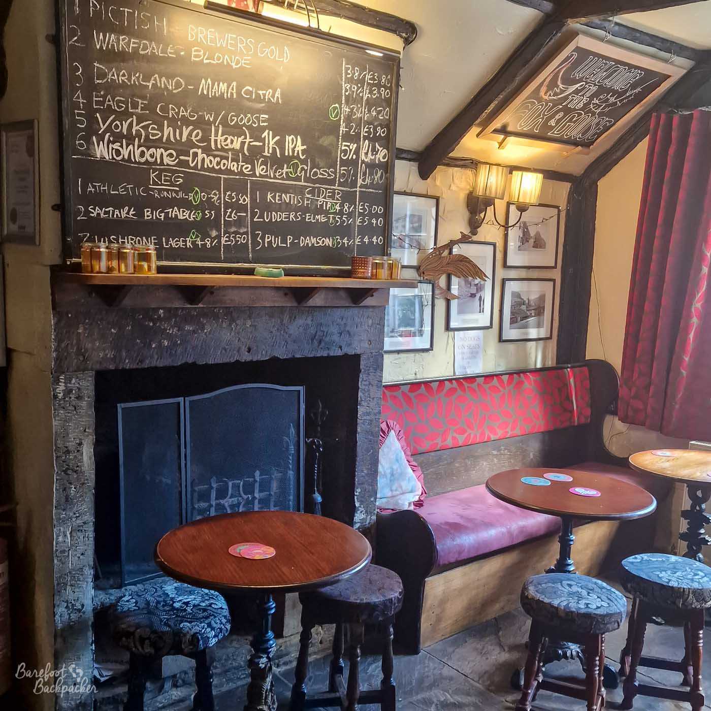 Cosy seating area inside the Fox and Goose in Hebden Bridge, centred around a large dark stone fireplace with a black fireguard. Small round wooden tables and mismatched upholstered stools sit beside a red patterned bench, beneath a large handwritten chalkboard listing six cask beers plus keg beers and ciders with their prices. Exposed dark wooden beams cross the low ceiling, framed pictures decorate the walls, and a hanging sign reads Welcome to the Fox & Goose.