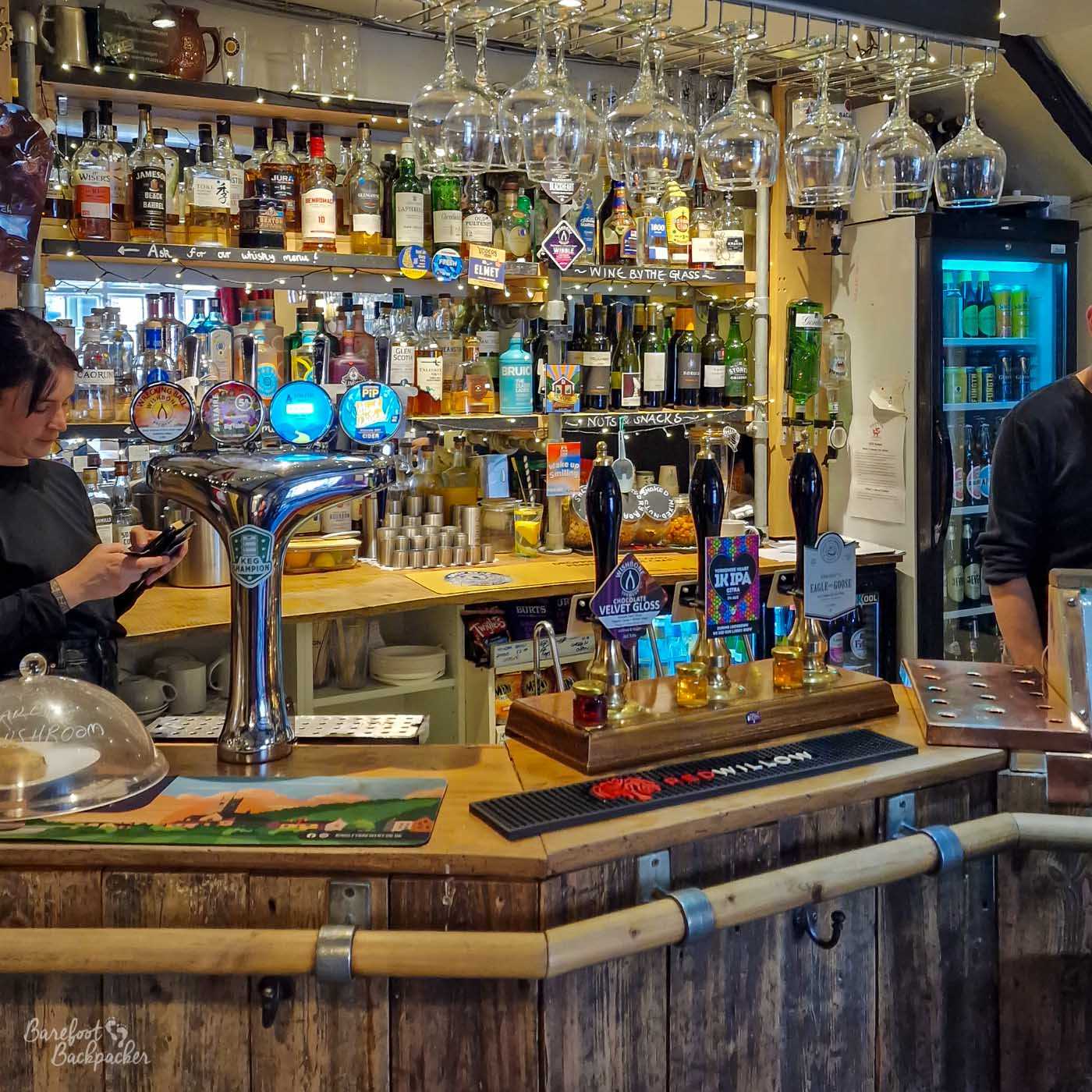 Close view of the compact, densely stocked bar inside the Fox and Goose in Hebden Bridge. Rows of whisky, gin, wine and other bottles fill illuminated wooden shelves behind several hand-pulls and keg taps, while large wine glasses hang upside down from racks overhead. Handwritten labels on the shelving advertise whisky, wine and snacks, and two members of staff stand partly visible at either side. The wood-panelled bar and crowded displays give the pub a cosy, traditional feel.