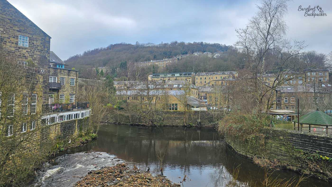 A broad section of dark canal water in Hebden Bridge, surrounded by old stone buildings climbing up the wooded valley sides. Bare winter trees line the banks, with a small weir or area of fast-flowing water in the foreground. The town rises densely behind the canal beneath an overcast grey sky.