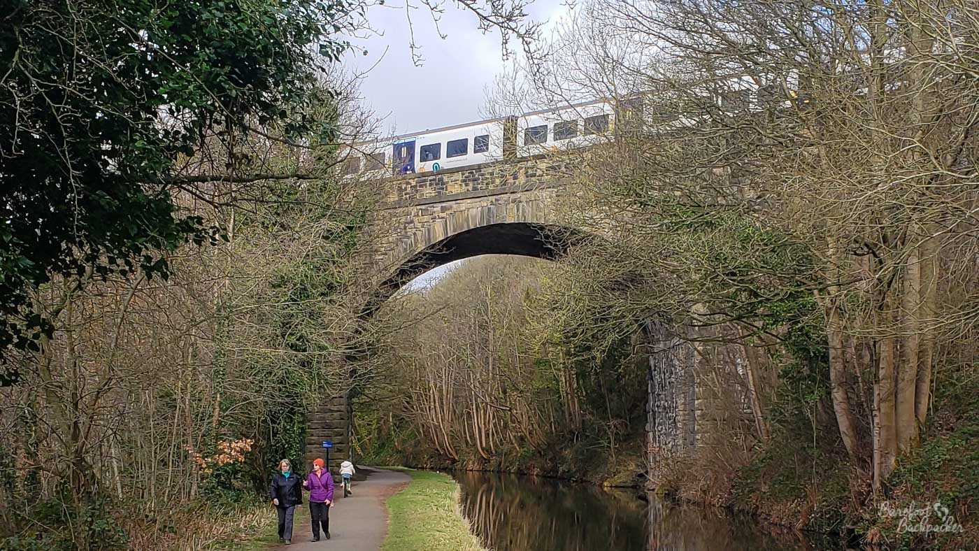 A tall stone railway viaduct crosses the canal through leafless woodland, with a passenger train travelling across the top. The large central arch spans both the narrow waterway and its paved towpath, where several people are walking below. Bare branches frame the bridge and partially obscure its stone piers.