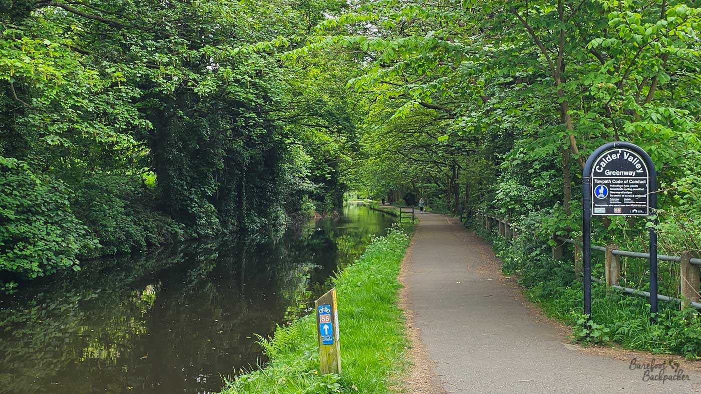 A paved towpath runs alongside a narrow canal through dense, leafy woodland. A black sign beside the path identifies the route as the Calder Valley Greenway, while a smaller blue and red cycle marker shows National Cycle Route 66. Trees arch over both the water and path, creating a thick green canopy.