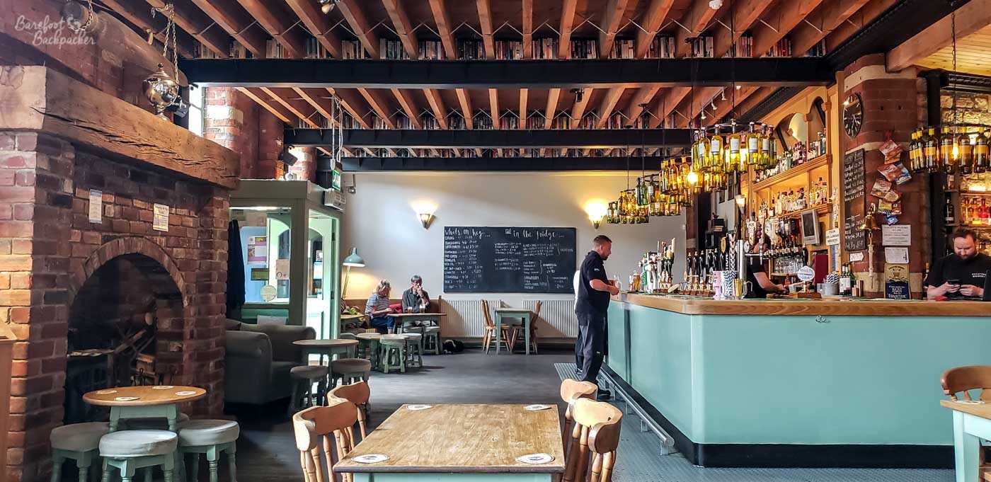 The spacious main bar combines exposed brickwork, timber beams and high ceilings with pale green woodwork and warm lighting suspended from bottles above the long wooden counter. Large communal tables and scattered armchairs fill the open floor, while a chalkboard behind the bar lists beers and ciders alongside shelves stocked with bottles and spirits. The former industrial building has been transformed into a bright, welcoming venue that still retains much of its original character.