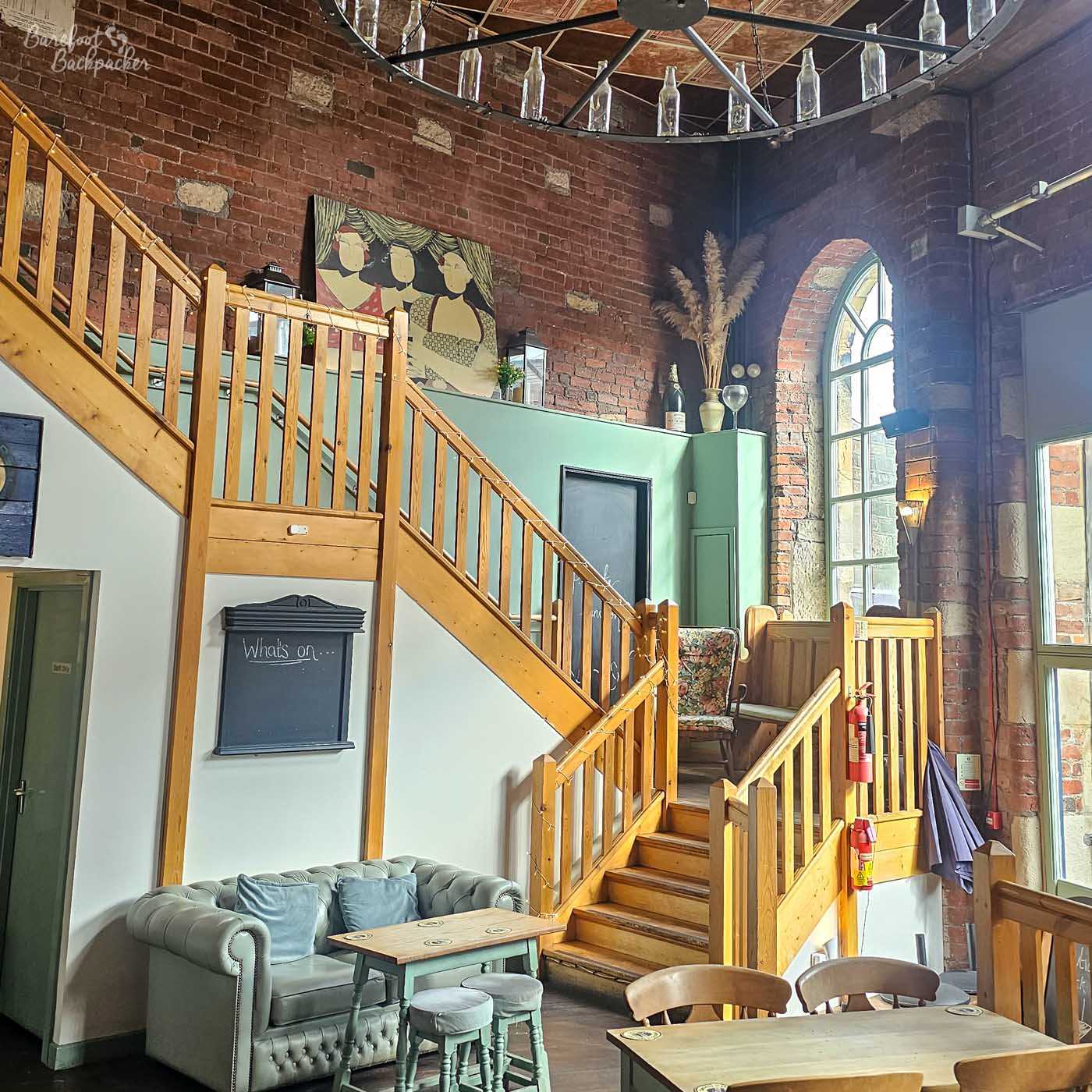 A broad wooden staircase rises through a lofty brick-lined hall beneath a circular metal chandelier decorated with empty glass bottles. Soft daylight pours through tall arched windows, illuminating pale green walls, exposed brick pillars and comfortable seating arranged around small wooden tables. The mixture of industrial architecture, reclaimed materials and contemporary furnishings gives the space an airy yet characterful feel.
