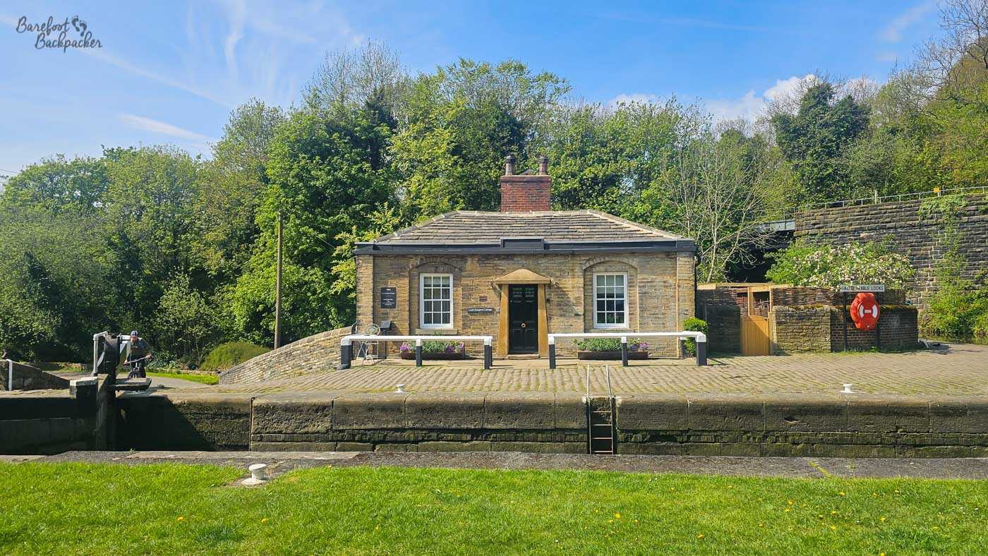 A small, symmetrical stone cottage stands immediately beside a canal lock, with two white-framed windows flanking its central black door. Black-and-white railings run along the paved lockside in front, with a ladder descending into the empty lock chamber. Trees rise densely behind the cottage, while a bright orange lifebuoy is mounted on the stone wall to the right.