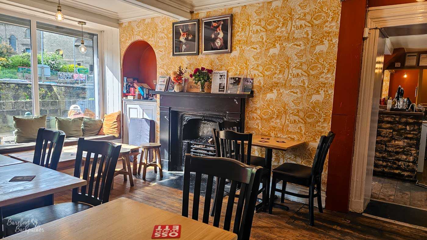 Inside The Fox, wooden tables and chairs surround a traditional black fireplace set against mustard-yellow patterned wallpaper. Framed fox illustrations hang above the mantelpiece, while sunlight pours through the large front window onto cushioned window seating, creating a warm and homely atmosphere.