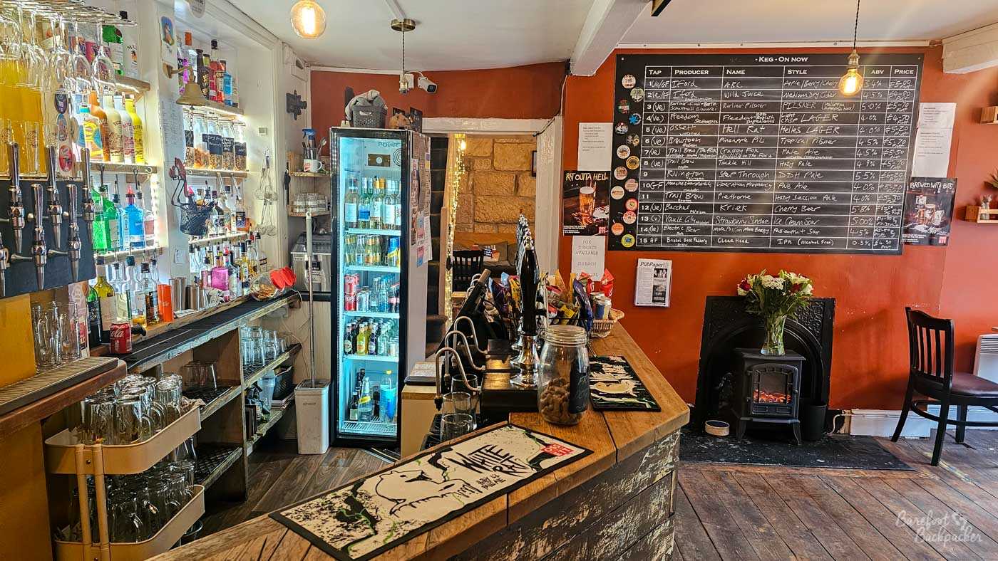 The compact wooden bar at The Fox displays rows of beer pumps beneath a large blackboard listing the current beers and ciders on tap. Shelves of spirits and bottled drinks line one wall, while a small stove and fireplace add warmth to the cosy room. The focus is clearly on the wide range of drinks available.