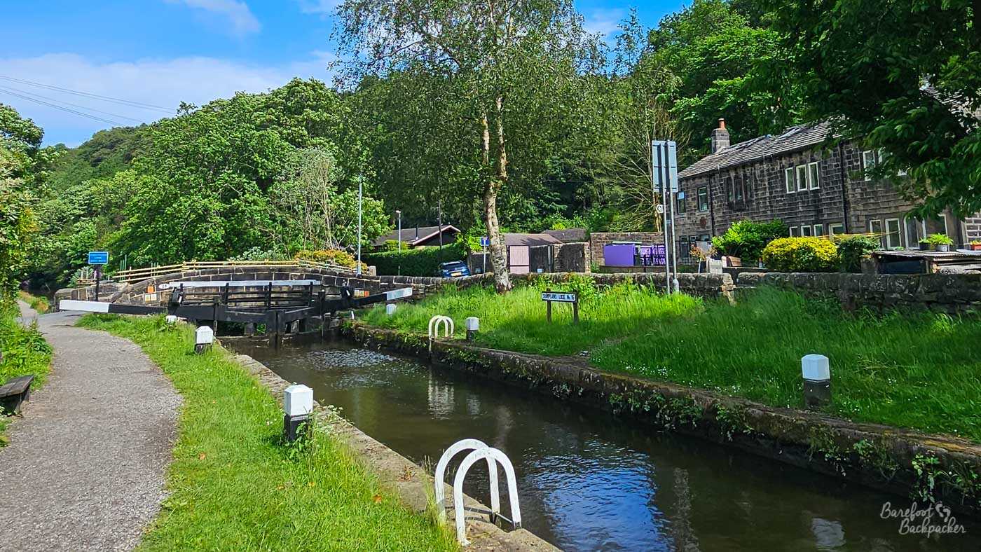 A canal lock in a leafy residential setting, with black-and-white lock gates ahead and a towpath running alongside the water. Traditional dark stone cottages stand behind a stone wall on the opposite bank, surrounded by mature trees and long summer grass. White-painted metal ladders descend into the canal at intervals along the lock.