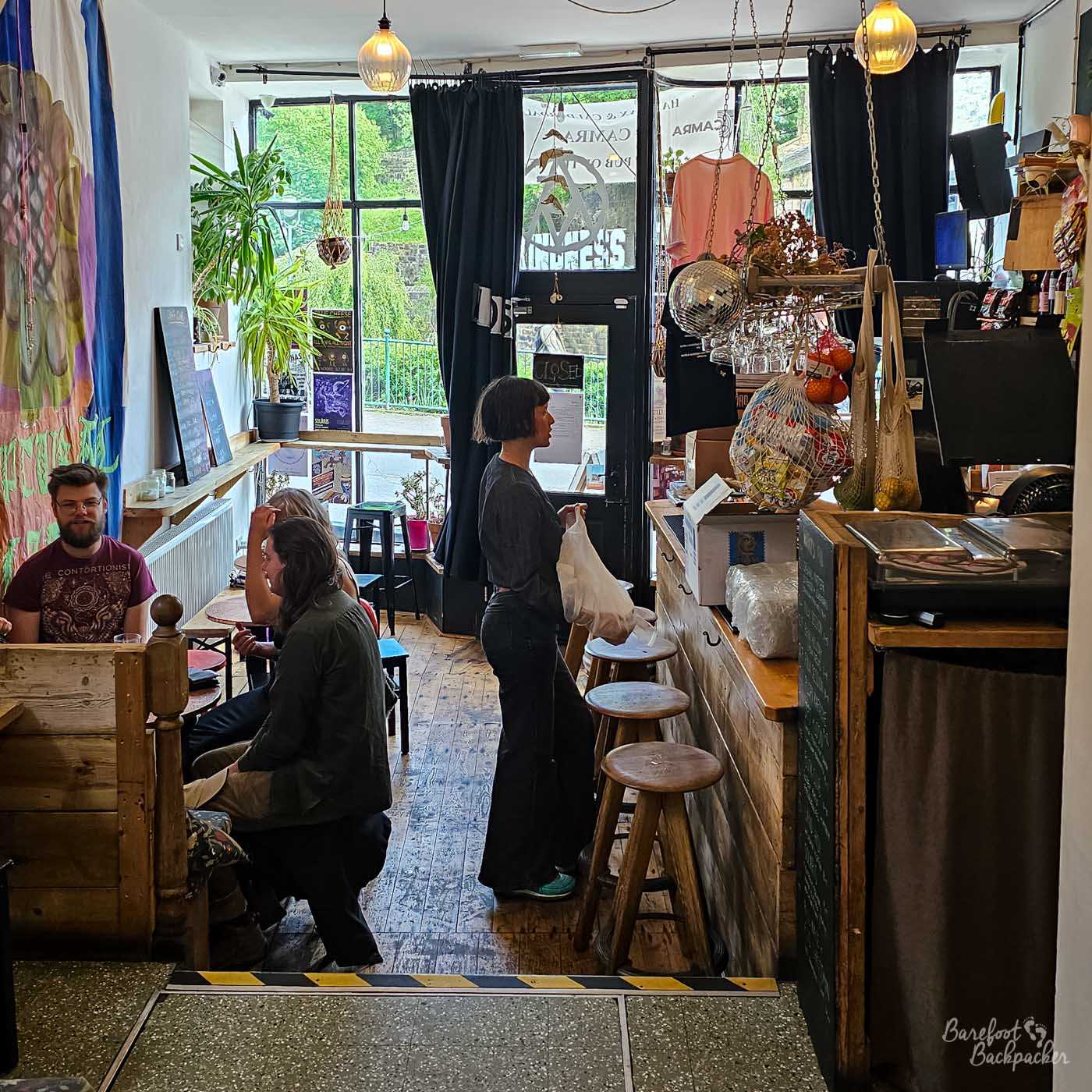 Looking from the rear of Nan Moor's towards the entrance, a customer stands at the bar while a small group chat at a wooden booth nearby. Large front windows let daylight flood into the narrow room, where plants, posters and eclectic decorations create a relaxed café-meets-bar atmosphere. The compact space feels busy, friendly and informal.