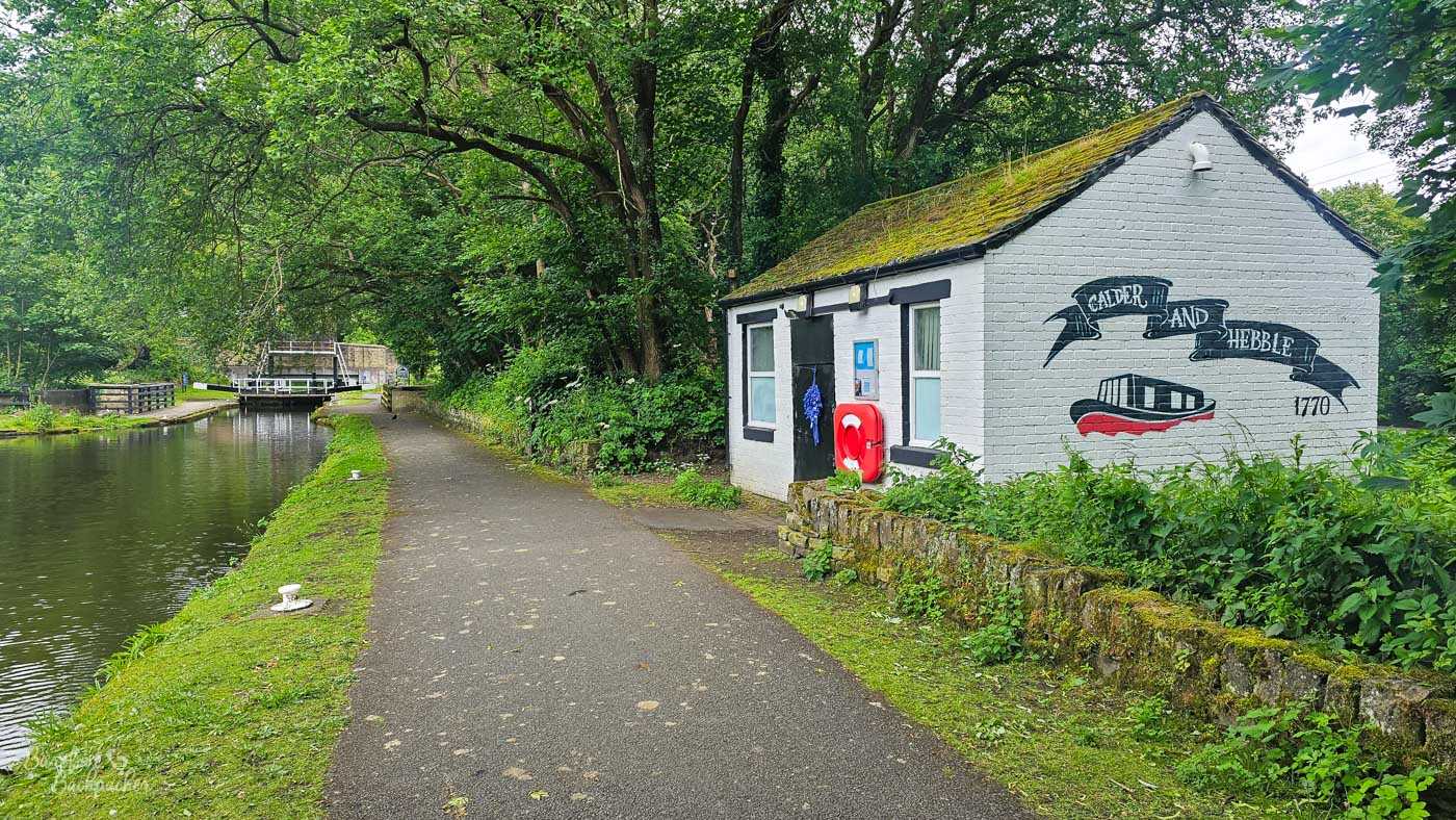A small white-painted brick building with a moss-covered pitched roof stands beside a wooded stretch of canal and towpath. A large painted mural on its gable reads Calder and Hebble 1770, surrounding an illustration of a traditional boat. A red lifebuoy hangs beside the building, with lock gates visible farther along the canal beneath the trees.