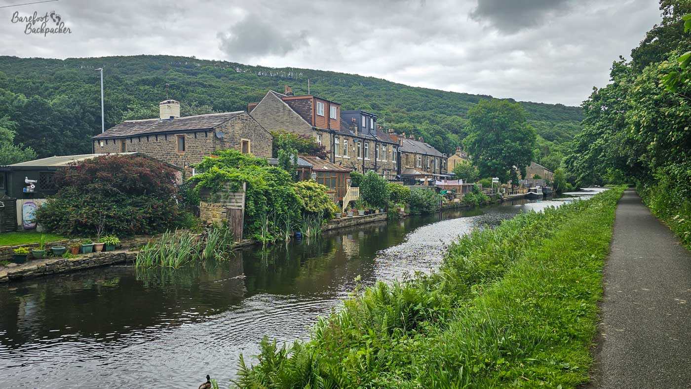 A broad canal runs beside a row of traditional stone houses and their waterside gardens, with a paved towpath continuing straight ahead on the opposite bank. Plants, sheds and small terraces crowd the canal edge beneath the houses, while densely wooded hills rise immediately behind the village. Heavy grey clouds hang above the green valley, giving the scene a distinctly damp Pennine feel.